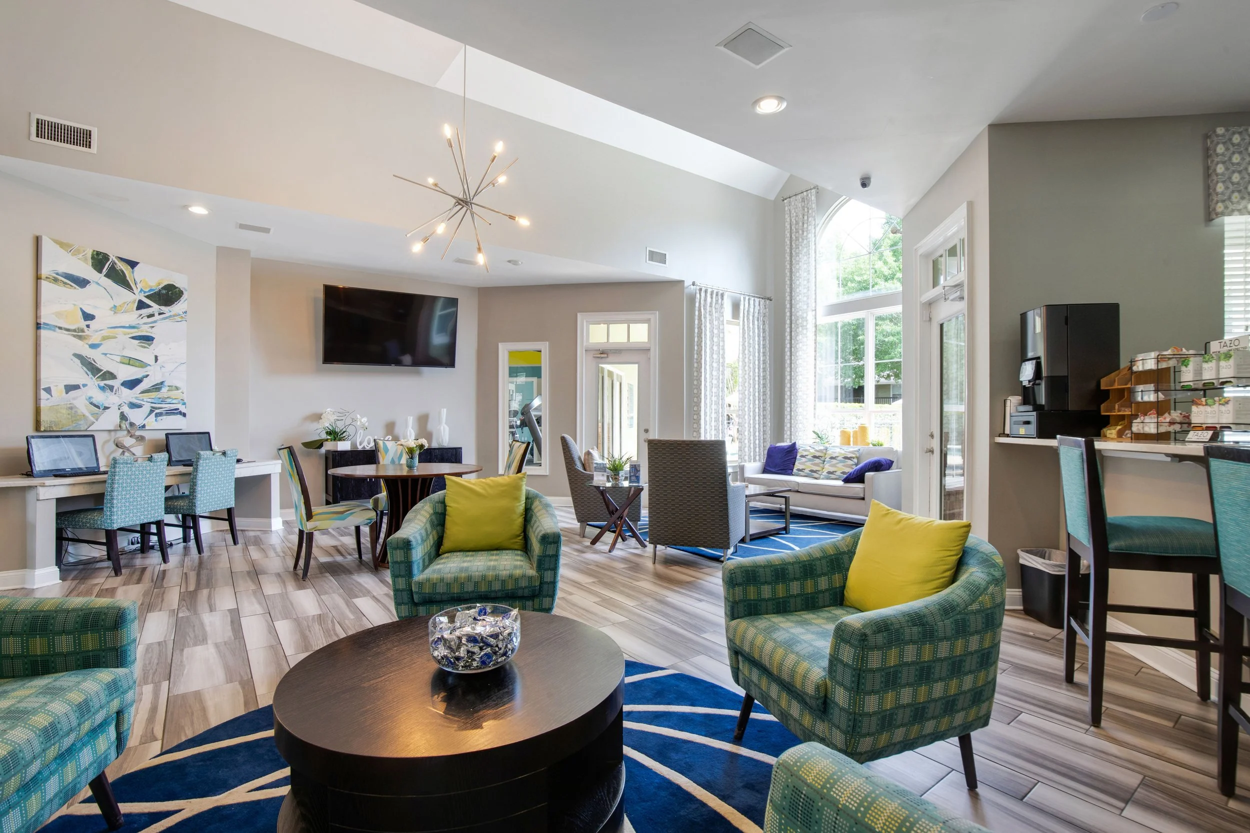 A bright and spacious lounge area with colorful armchairs, a round black coffee table with decorative glass bowl, modern artwork, large windows with curtains, a wall-mounted television, and a bar area with a coffee machine and snacks.