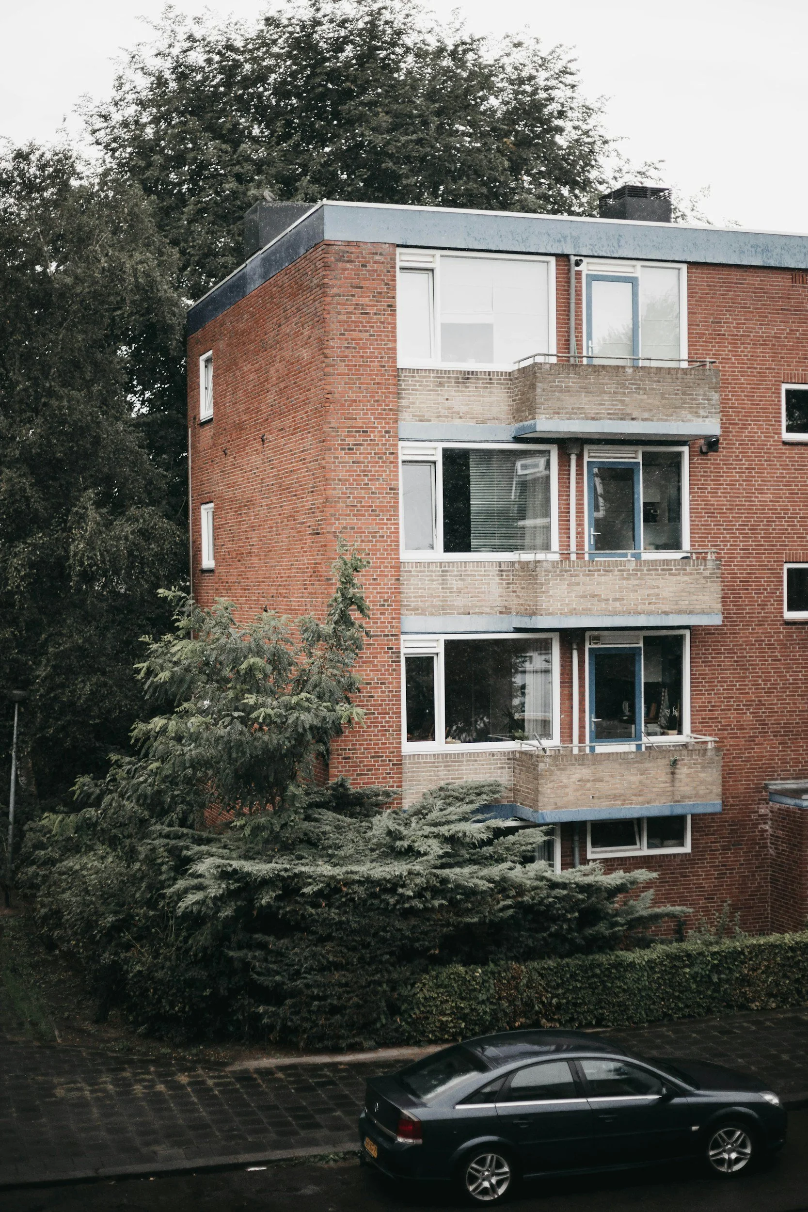 Multi-story brick apartment building with balconies, surrounded by trees and parked car in the foreground.