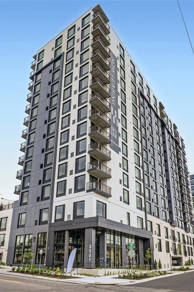 Modern multi-story residential building with balconies and large windows, located at 2501 on a city street, with trees and street signs in the foreground.