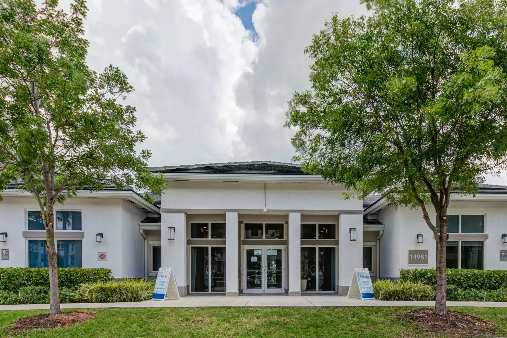 Front view of a modern office building with glass doors, surrounded by green trees and bushes, with signs and address number 14981 displayed.