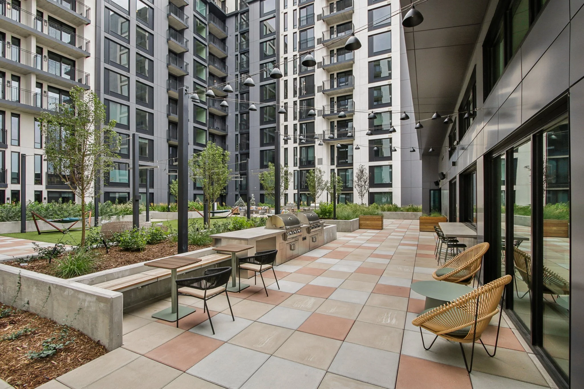 Modern outdoor patio area with seating, tables, grills, surrounded by tall apartment buildings and greenery.