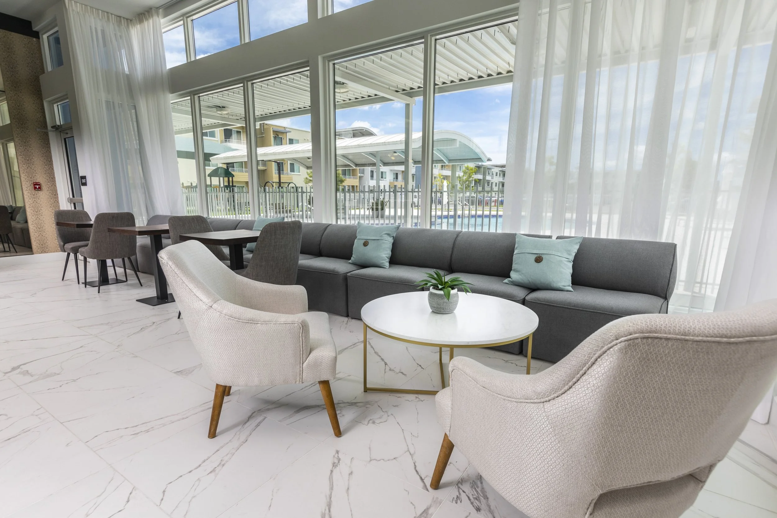 Indoor lounge area with beige and gray chairs, a gray sofa with light blue pillows, a white marble coffee table with a potted plant, and large windows with white curtains showing a pool and modern buildings outside.