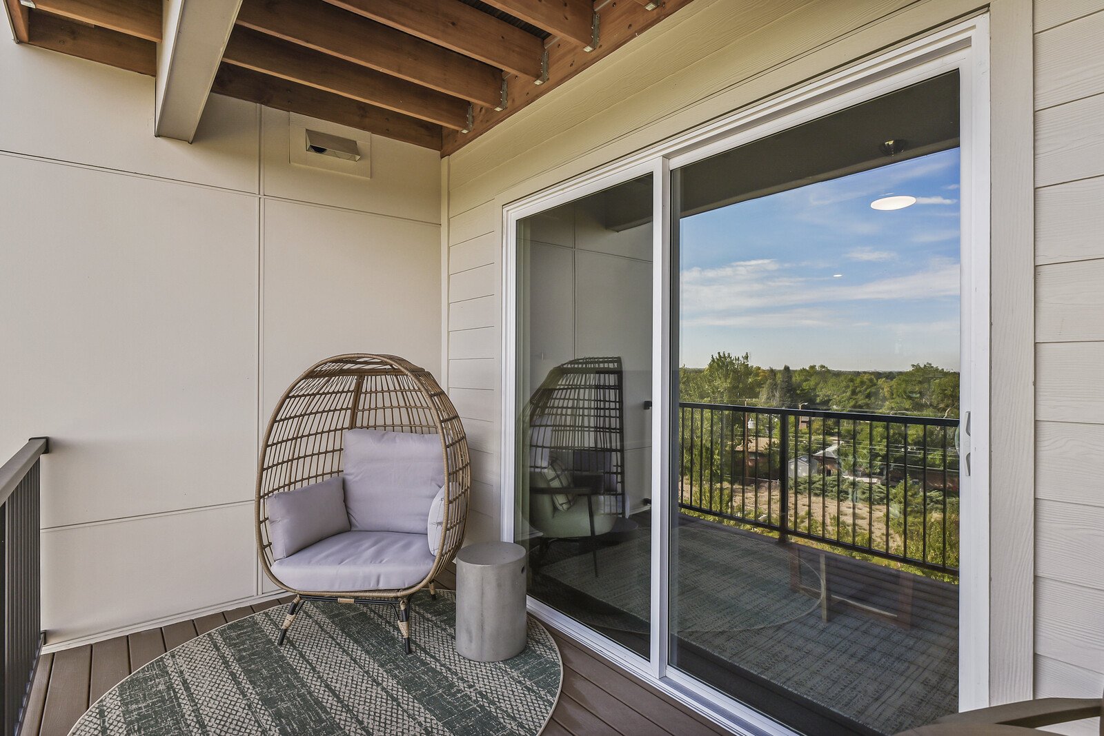 Balcony with wicker egg-shaped chair with cushions, small round concrete table, sliding glass door, view of green trees, blue sky, and nice weather.