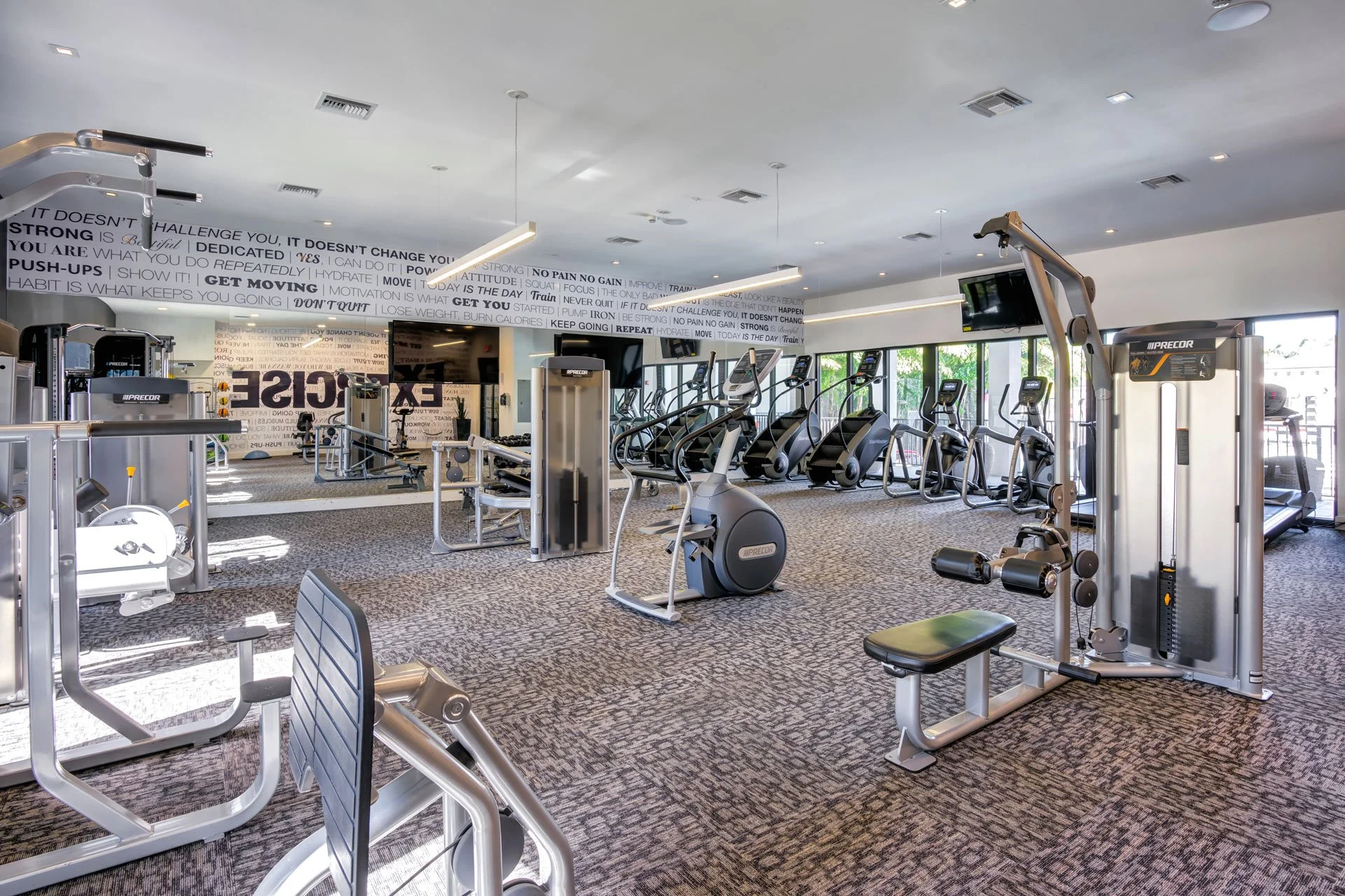 Empty gym with exercise machines including ellipticals, treadmills, and strength training equipment, with large windows showing outside greenery and Motivational quotes on the wall.