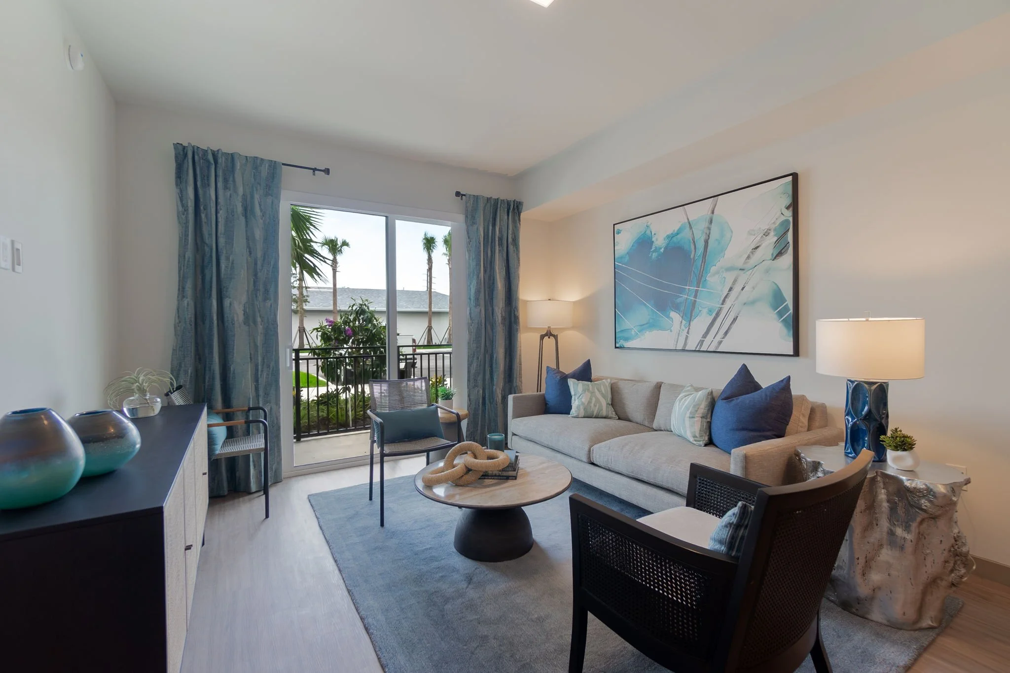 Living room with beige sofa, blue and white pillows, abstract blue artwork on the wall, sliding glass door with blue curtains, outdoor balcony with plants, mid-century modern coffee table, black chairs with cane backs, table with decorative objects, 