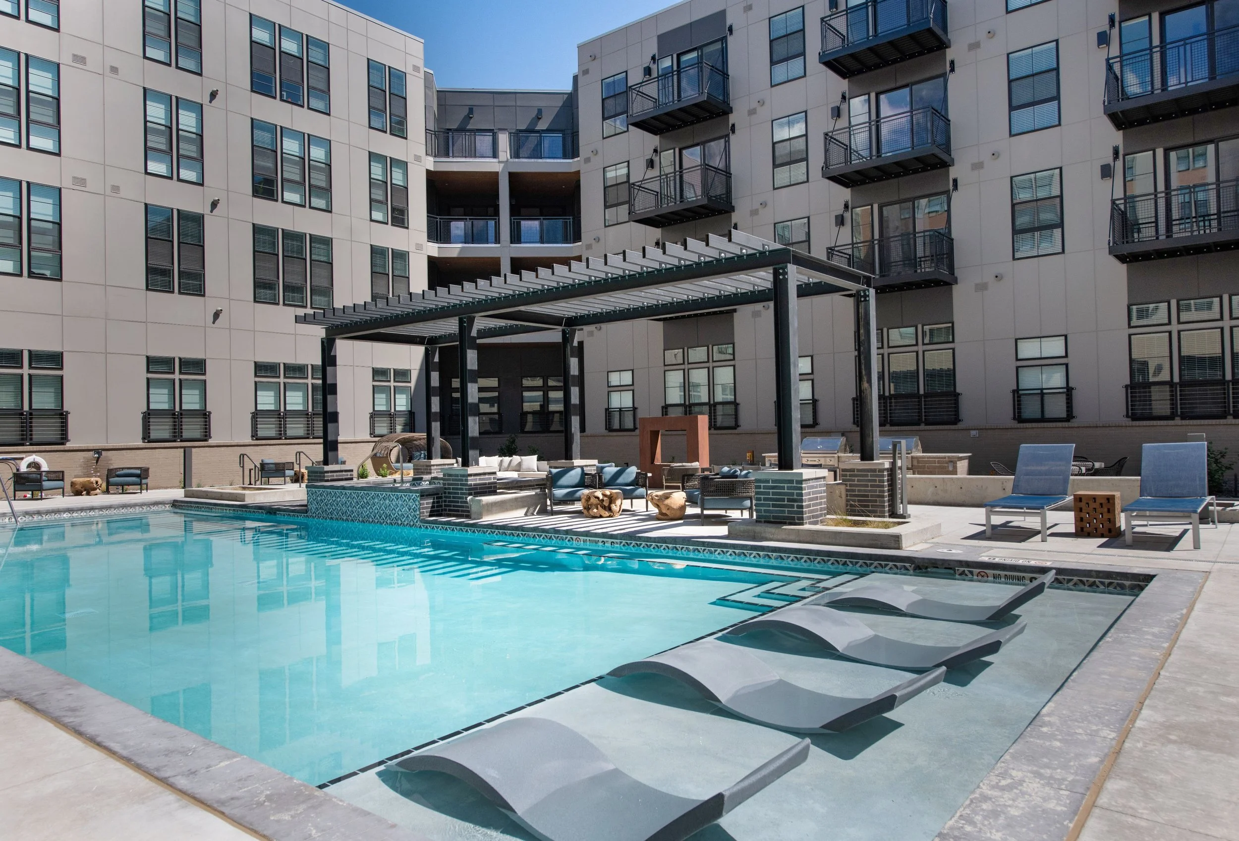 Outdoor swimming pool with three lounge chairs, a seating area with pillows and tables, and a pergola, surrounded by an apartment complex with multiple balconies.