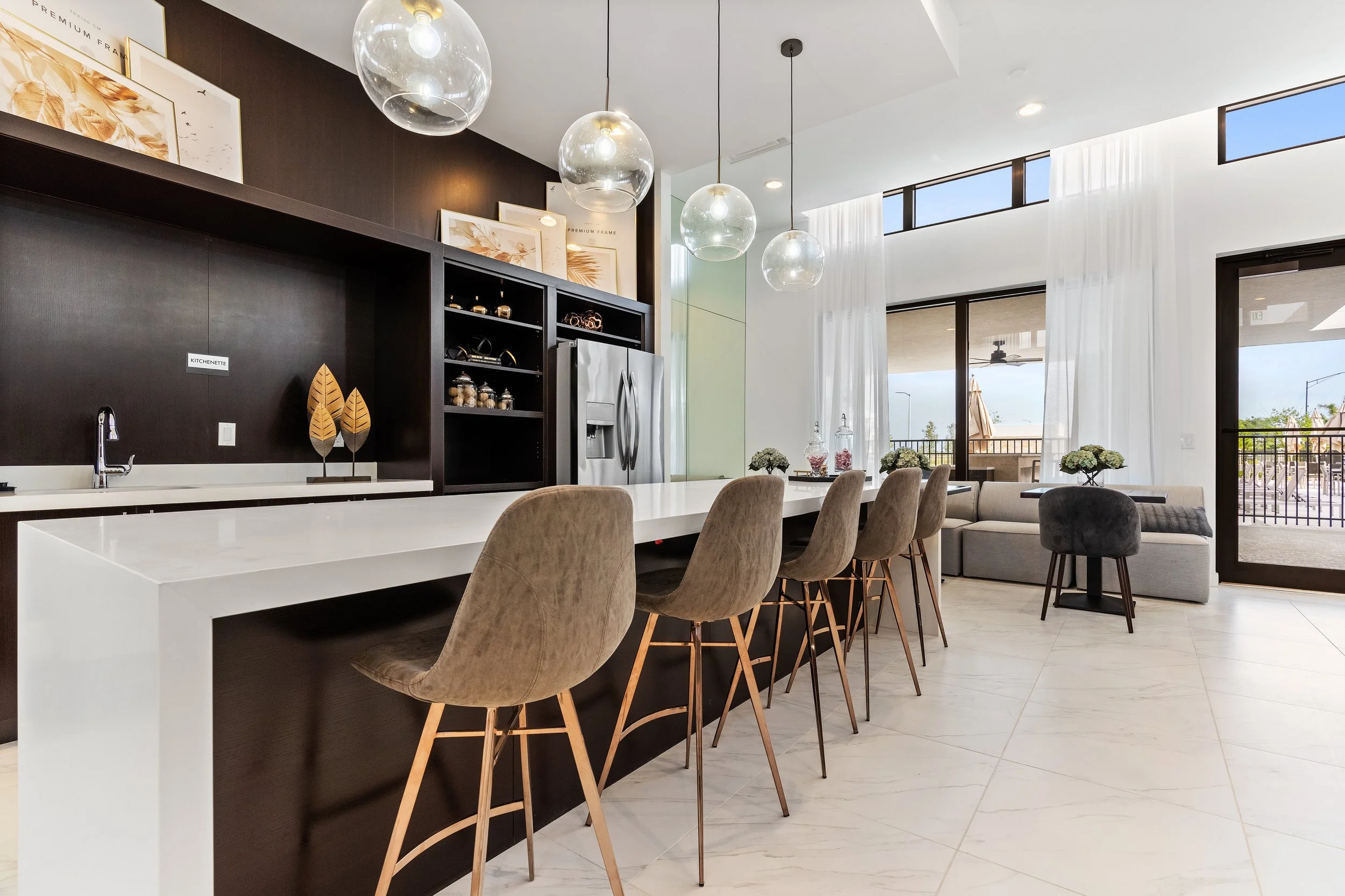 Modern kitchen and living area with white island, beige bar stools, black cabinetry, stainless steel refrigerator, glass pendant lights, white curtains, and seating area by sliding glass doors.