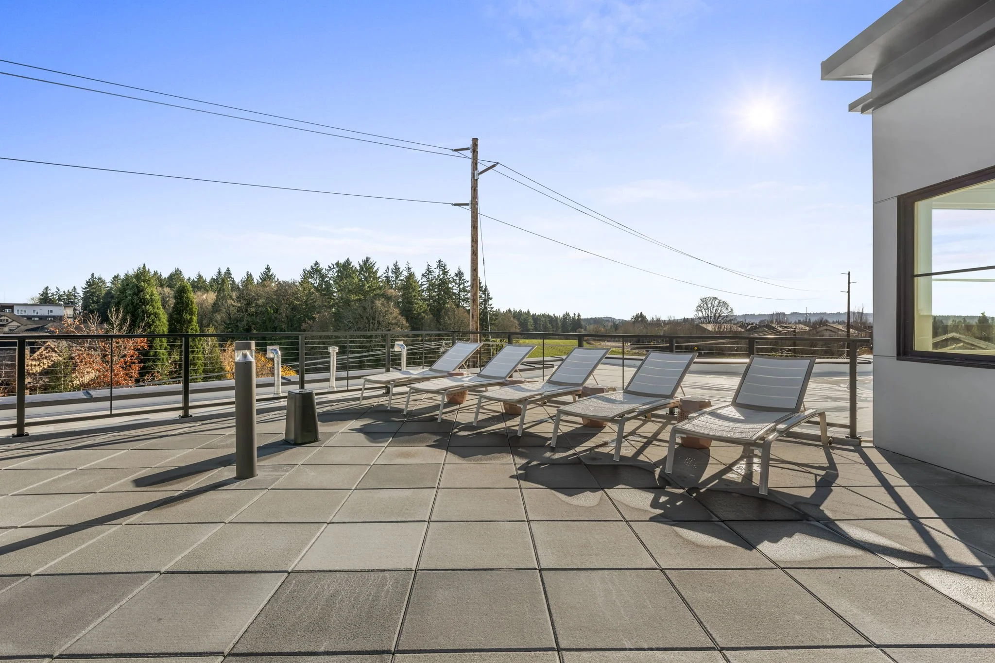 Empty rooftop patio with five lounge chairs facing a railing, overlooking trees and a clear sky with the sun shining.