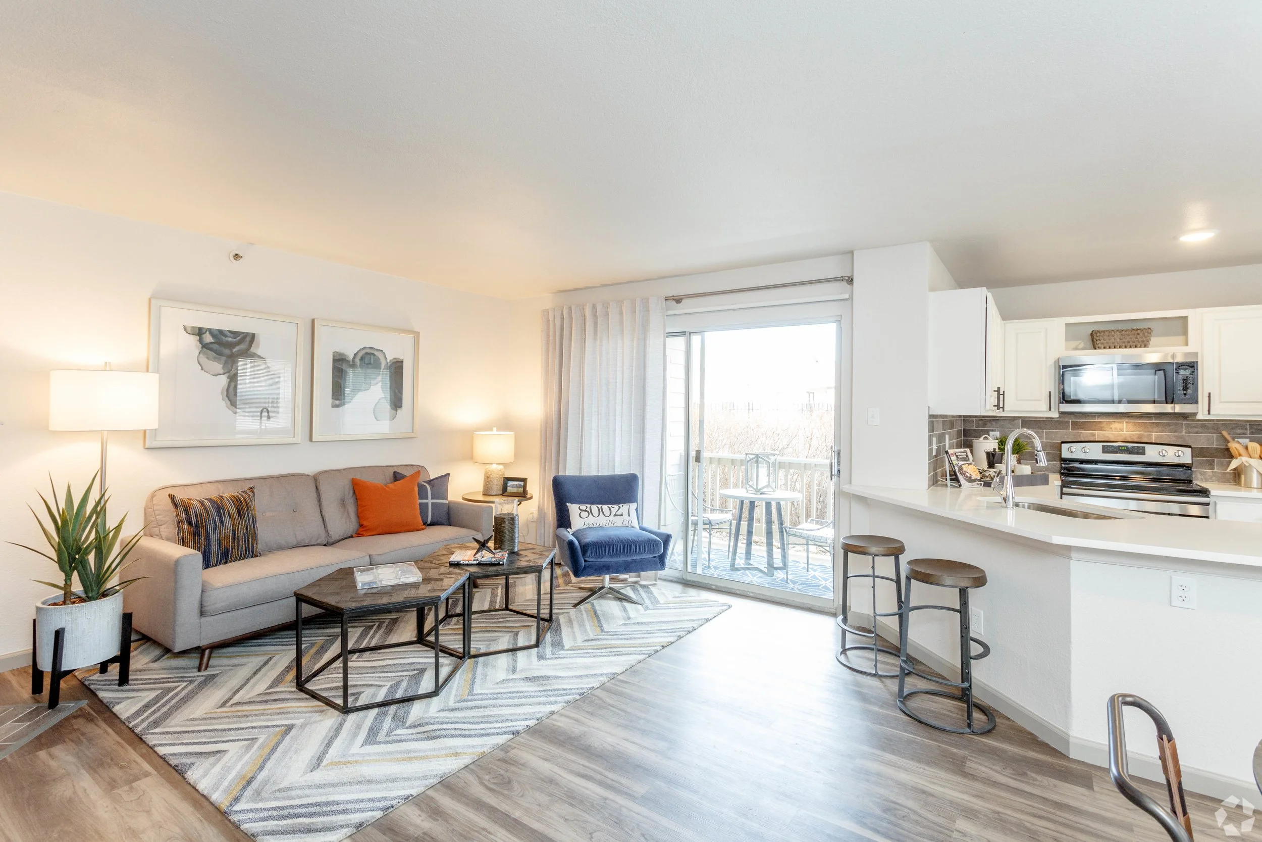 Living room with beige sofa, orange and blue cushions, abstract artwork above, side tables with lamps, a blue armchair near sliding glass door leading to balcony, kitchen with white cabinets, stainless steel microwave, and stove, barstools at kitchen