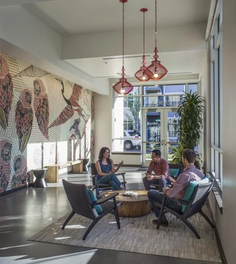 Three people sitting and talking in a modern bright lounge with a colorful bird mural, large window, and hanging red pendant lights.