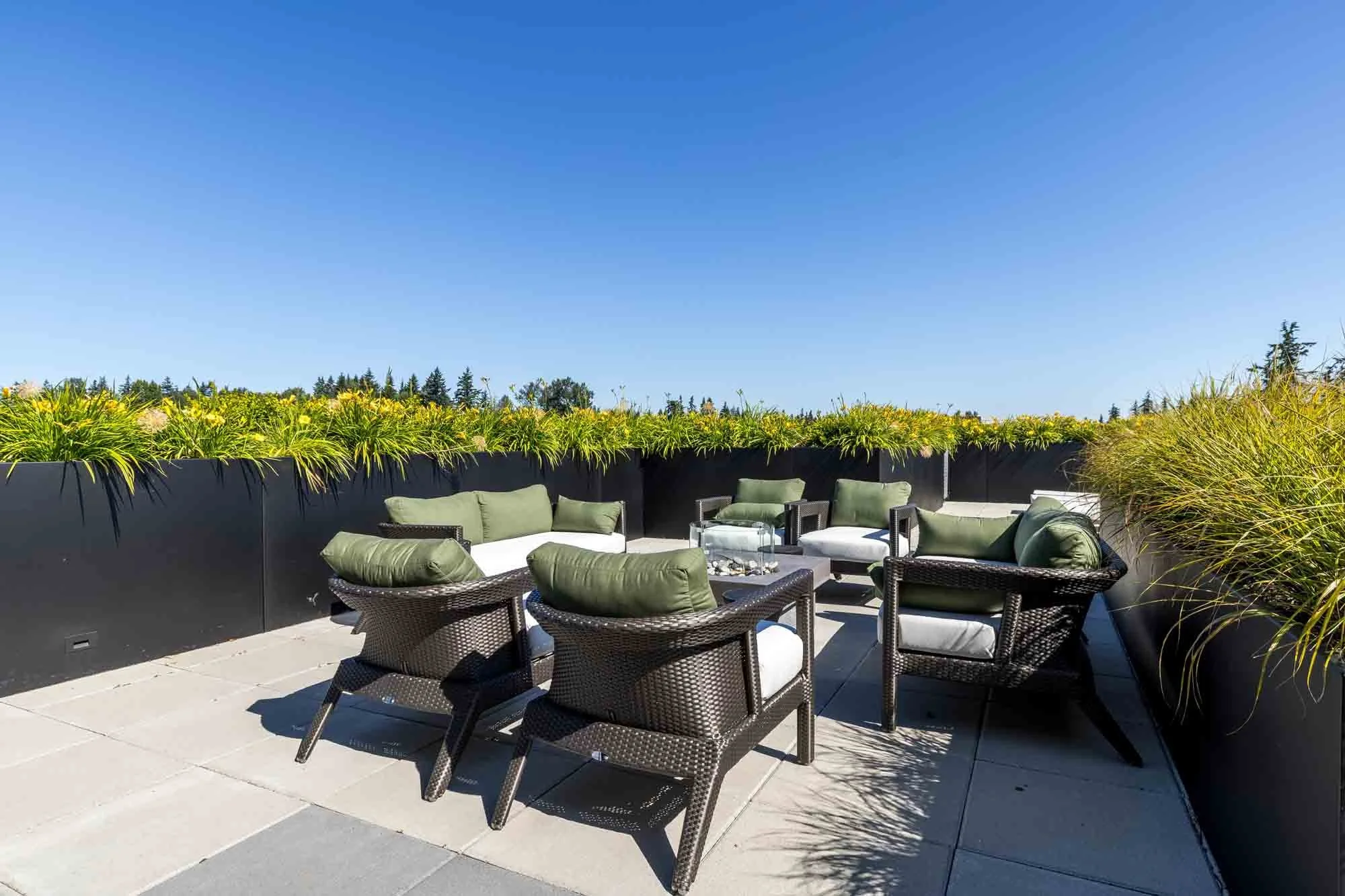 Rooftop patio with outdoor seating including wicker chairs with green cushions, a low table with a glass top, and a surrounding black planter filled with greenery, under a clear blue sky.