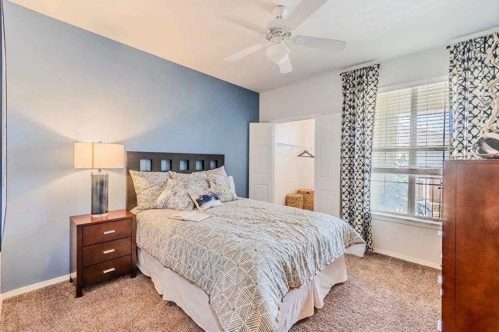 A bedroom with a king-sized bed, blue accent wall, bedside table with lamp, window with patterned curtains, and a closet with open door showing baskets inside.