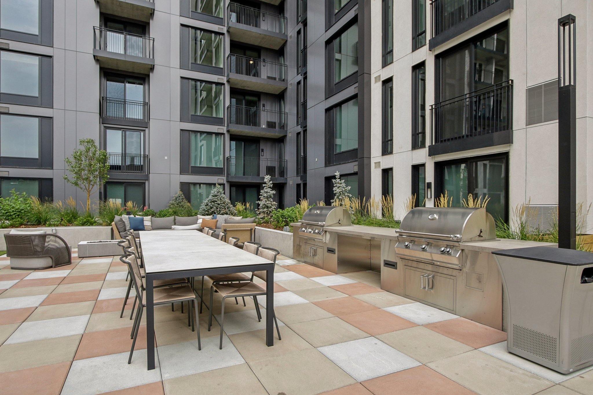 Outdoor apartment courtyard featuring a dining table with chairs, grilling stations, seating area, and landscaping, surrounded by a modern multi-story residential building with balconies.
