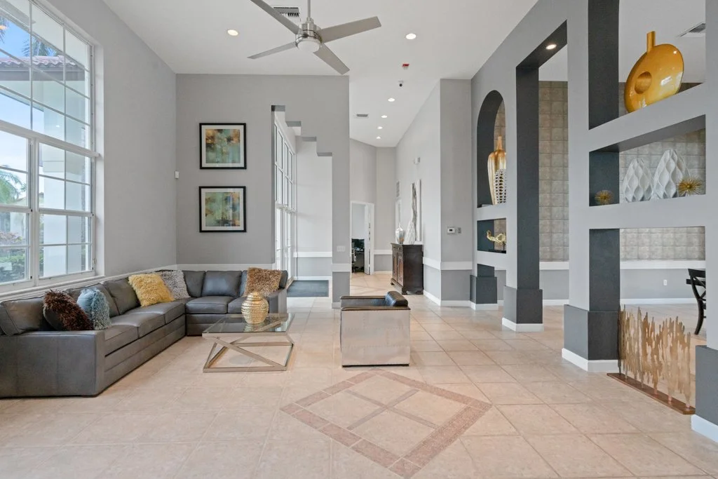 Modern living room with gray sectional sofa, glass coffee table, beige tiled floor, decorative pillows, large windows, gray and white walls with artwork, ceiling fan, open shelving with vases, and a tiled accent wall in the background.