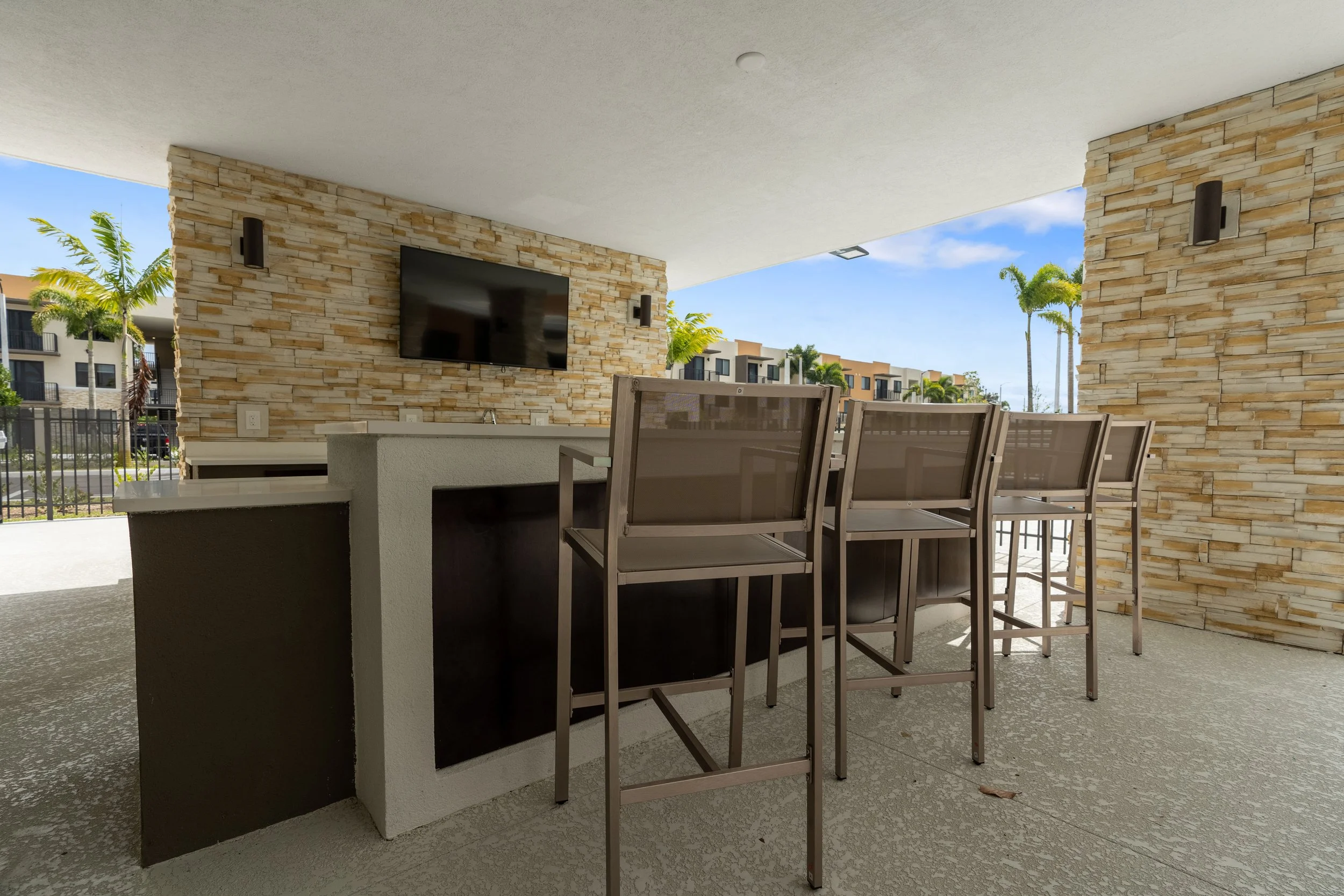 Outdoor patio with bar seating, a mounted TV on stone wall, and blue sky with some clouds and palm trees in the background.
