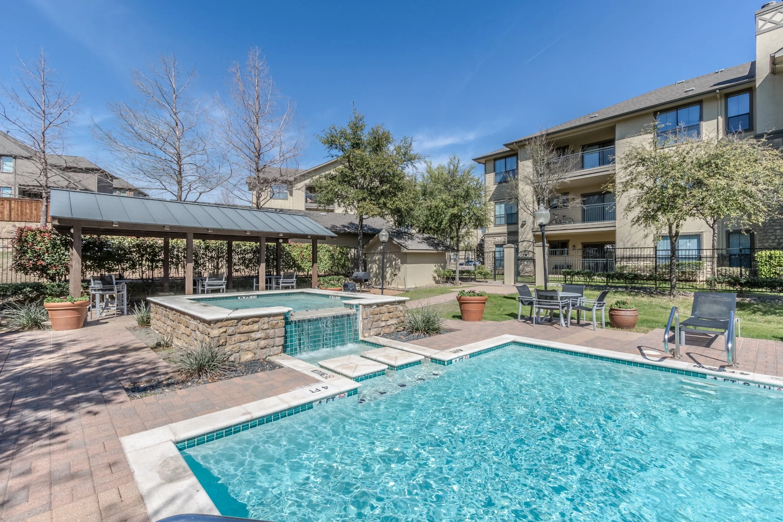 Outdoor swimming pool area in an apartment complex with chairs, tables, potted plants, trees, and a covered seating area, under a clear blue sky.