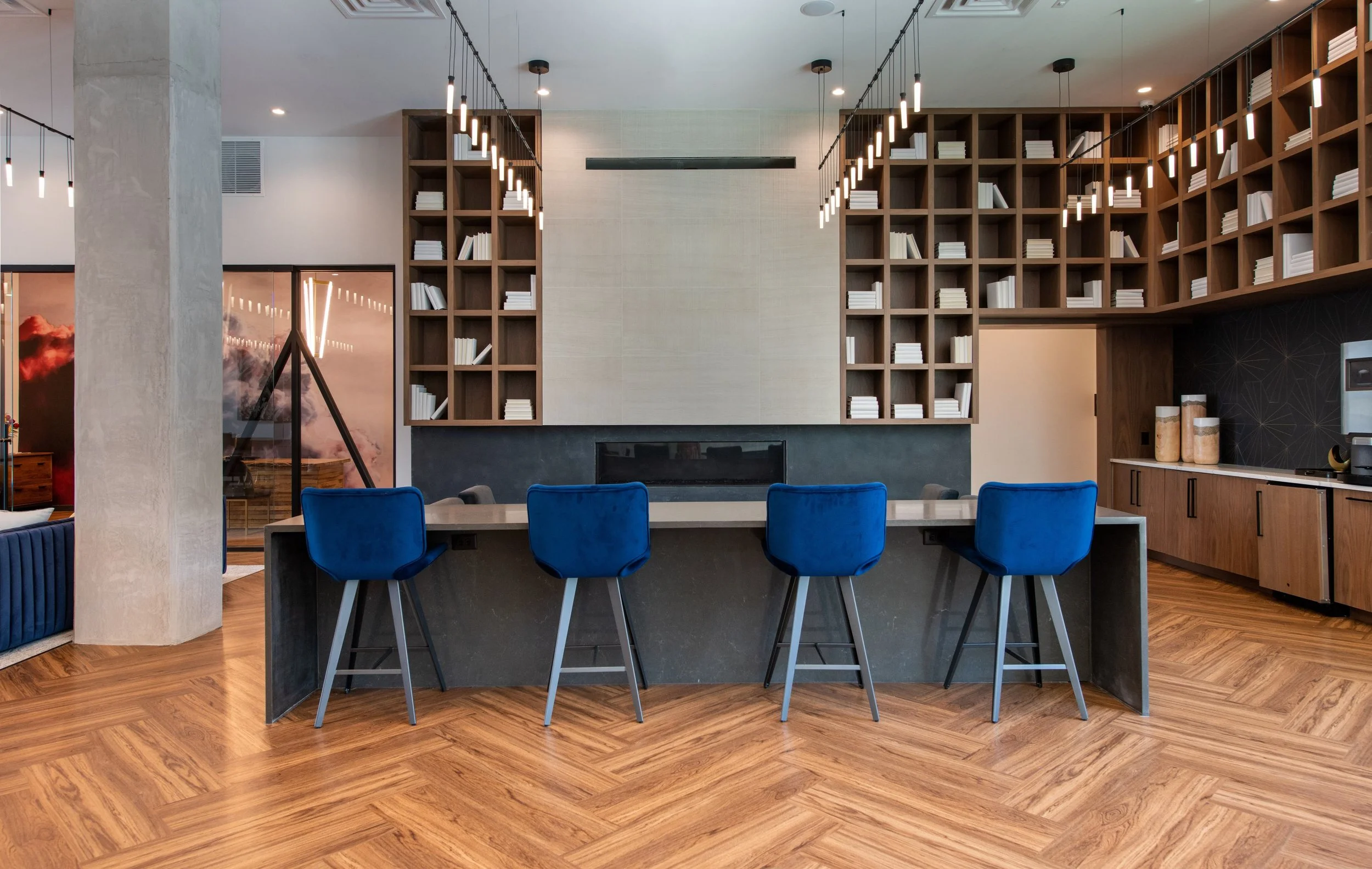 Modern indoor lounge area with a long island counter and four blue chairs. Behind the counter, a large wall features built-in wooden bookshelves filled with white books. The room has wooden flooring and contemporary hanging light fixtures.