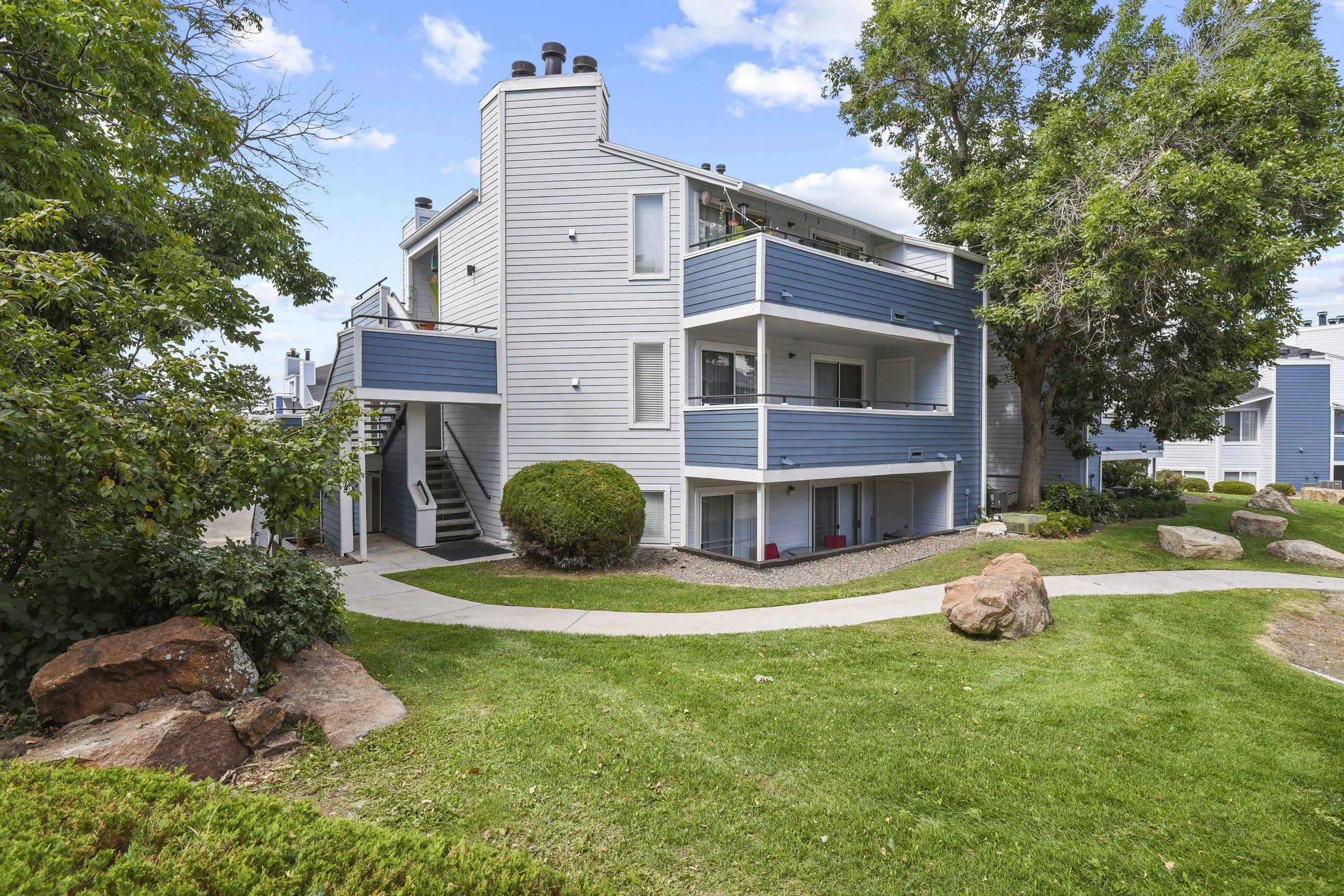A three-story residential apartment building with blue and white siding, balconies, staircases, and surrounding green landscape with rocks and trees.