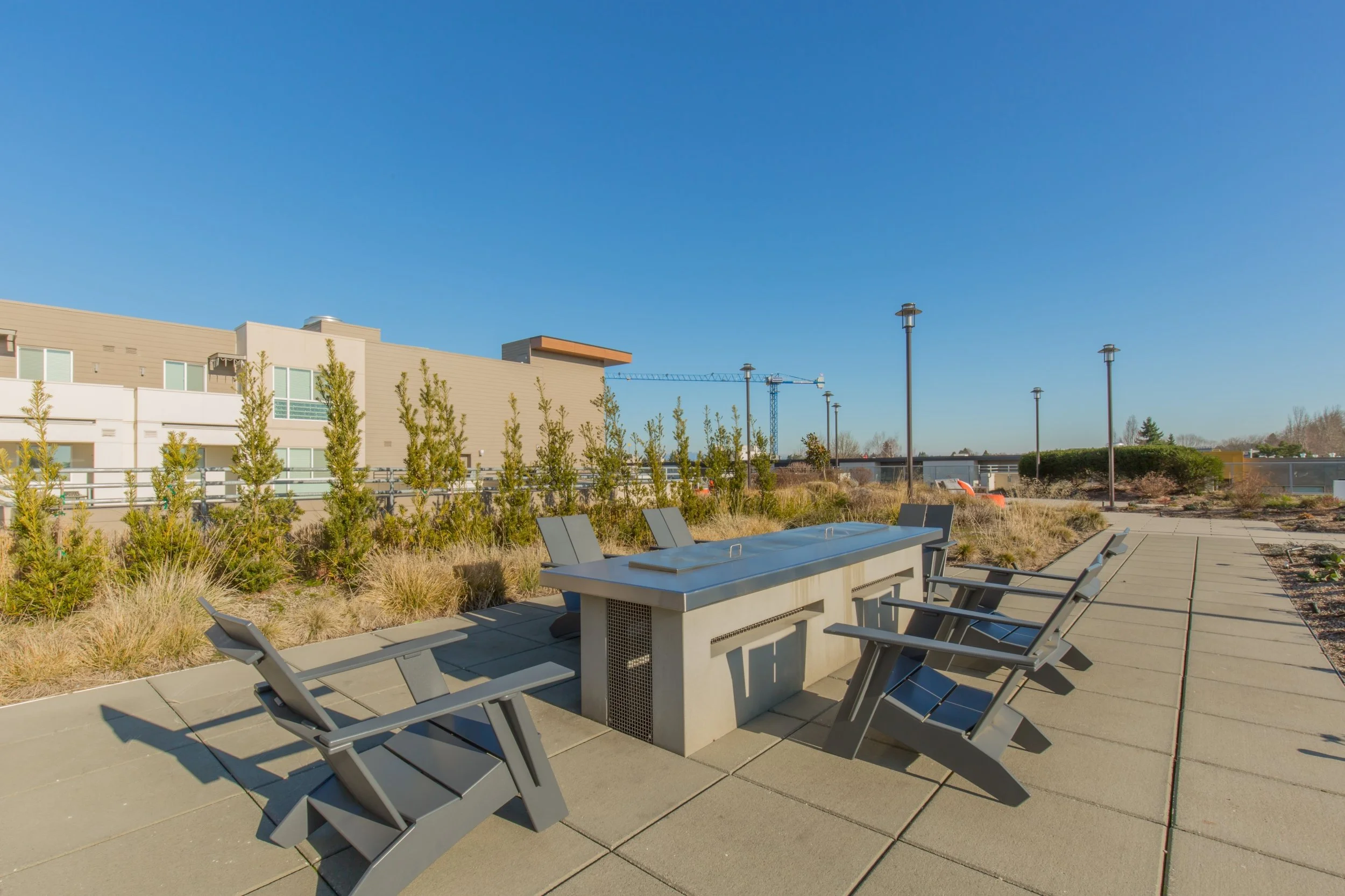 Outdoor seating area with a metal table and six chairs under a clear blue sky, surrounded by landscaped plants and shrubs, with modern buildings and construction crane in the background.