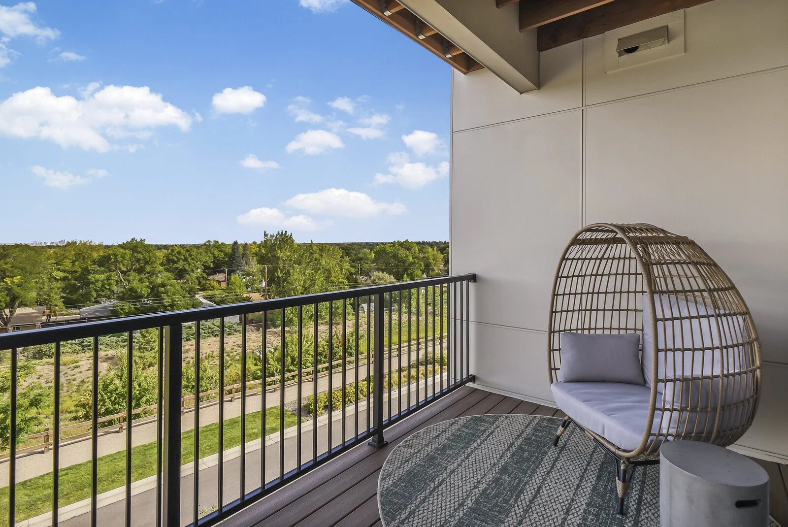 Balcony with outdoor chair with cushion, small side table, and view of green trees, grass, and a clear blue sky with clouds.