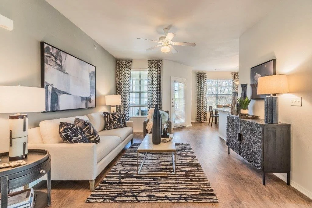 Living room with white sofa, black and white pillows, wooden floor, patterned curtains, windows, and a dining area in the background.