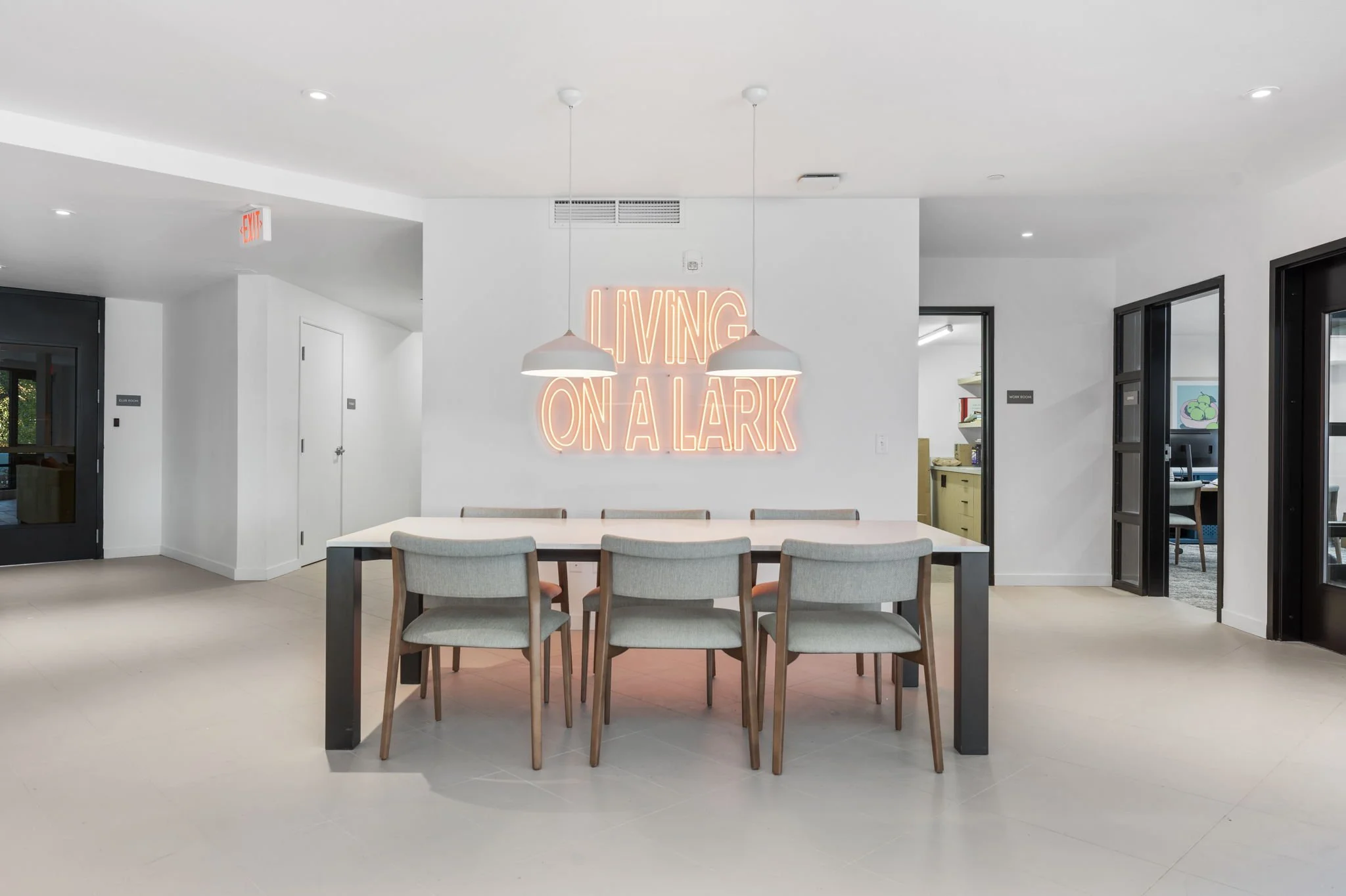 Modern dining room with a white table, six upholstered chairs, neon sign reading 'LIVING ON A LARK' on white wall, two hanging pendant lights, and glass doors leading to other rooms.