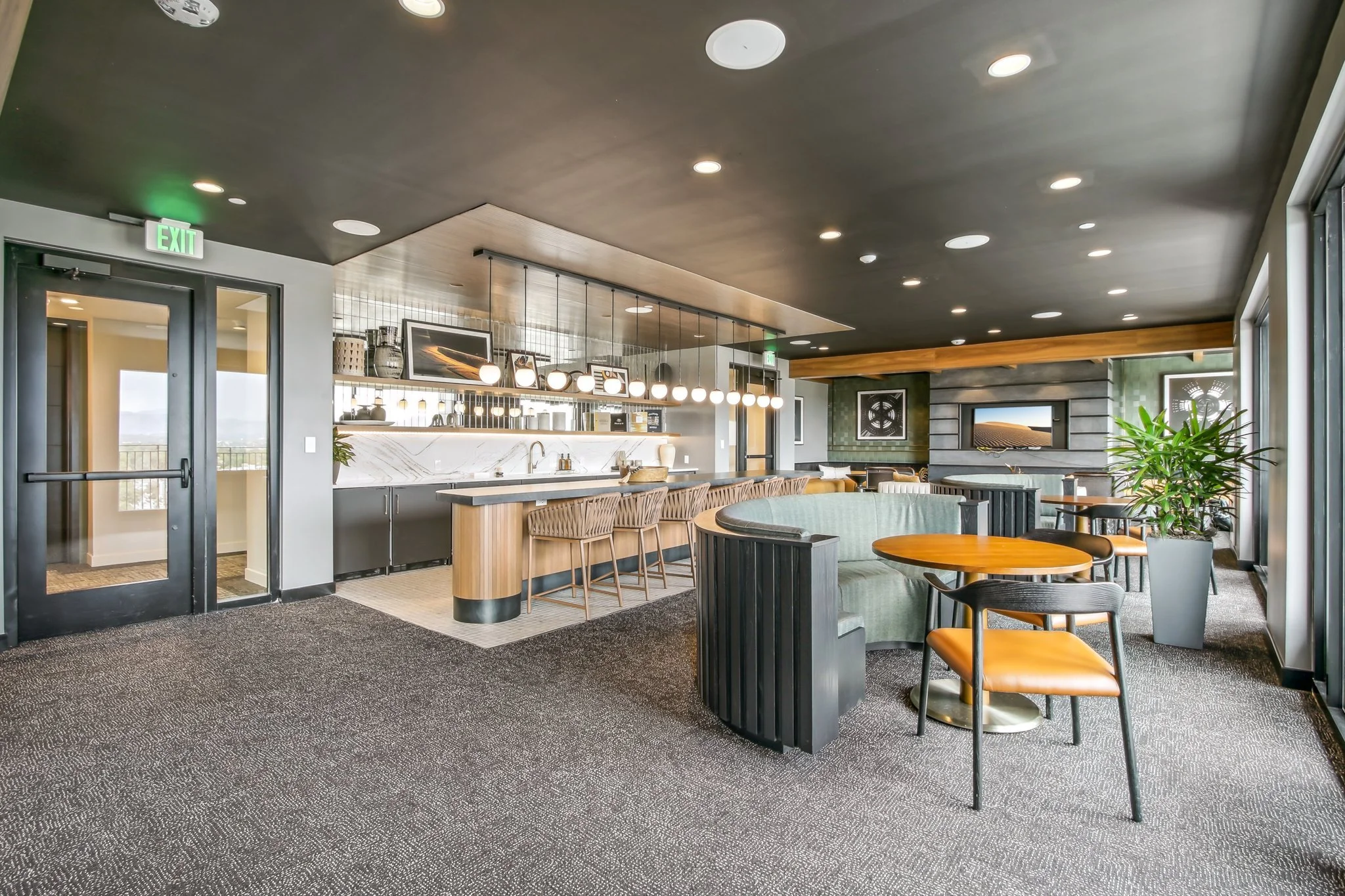Modern lounge area with a bar, seating, and a television, featuring a dark ceiling with recessed lighting and large windows with natural light.