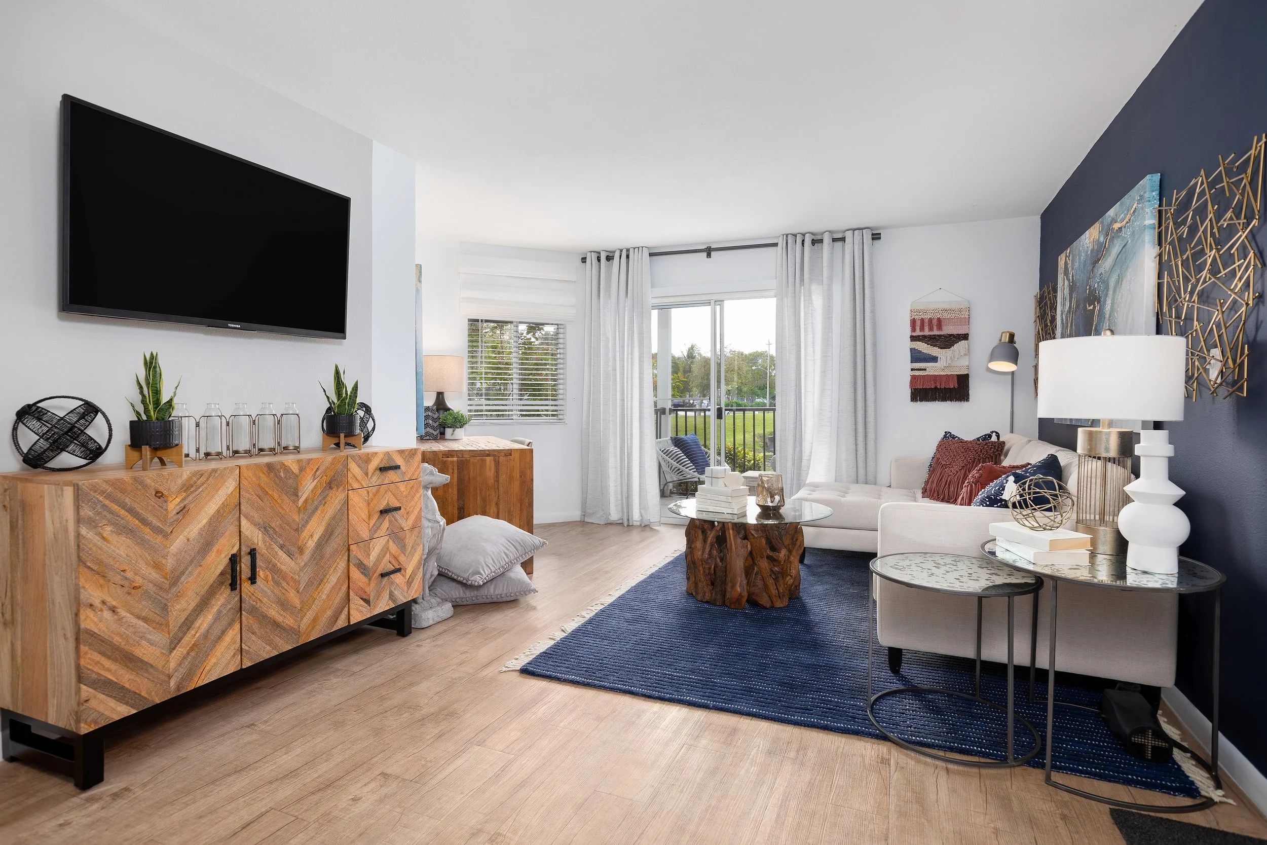 A modern living room featuring a wooden sideboard, a wall-mounted TV, a white sofa with red and navy cushions, a glass coffee table with a wooden base, a navy rug, a sliding glass door with white curtains, and various decorative items and artwork on 