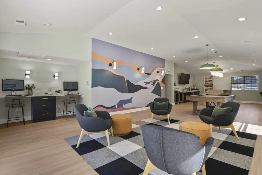 A spacious lounge area with modern seating, including four black armchairs with green cushions, and yellow ottomans. A checkered rug is on the wooden floor. The room features a large abstract mural on the wall, multiple wall-mounted light fixtures, a
