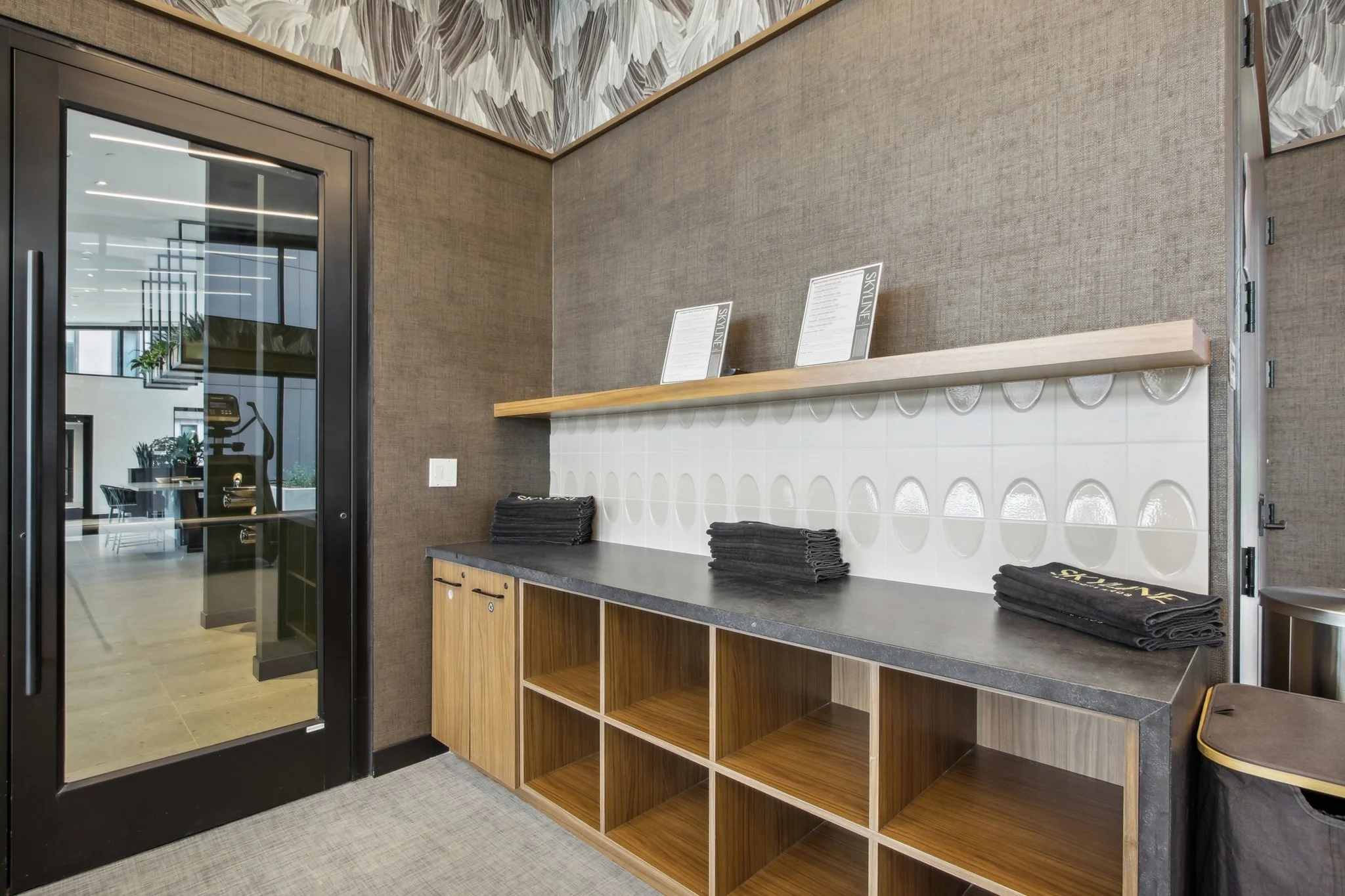 Indoor view of a reception or service area with a wooden cabinet, black countertop, and white tiled backsplash. Several stacks of black towels and two informational menus are on a wooden shelf. There is a glass door to the left revealing a fitness ro