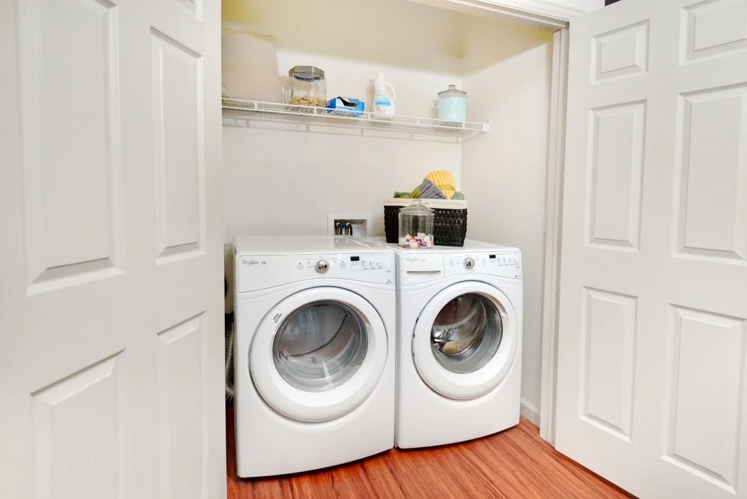 A small laundry room with white washer and dryer side by side, with open white double doors, a wire shelf above holding various items, and a black basket on top of the appliances.