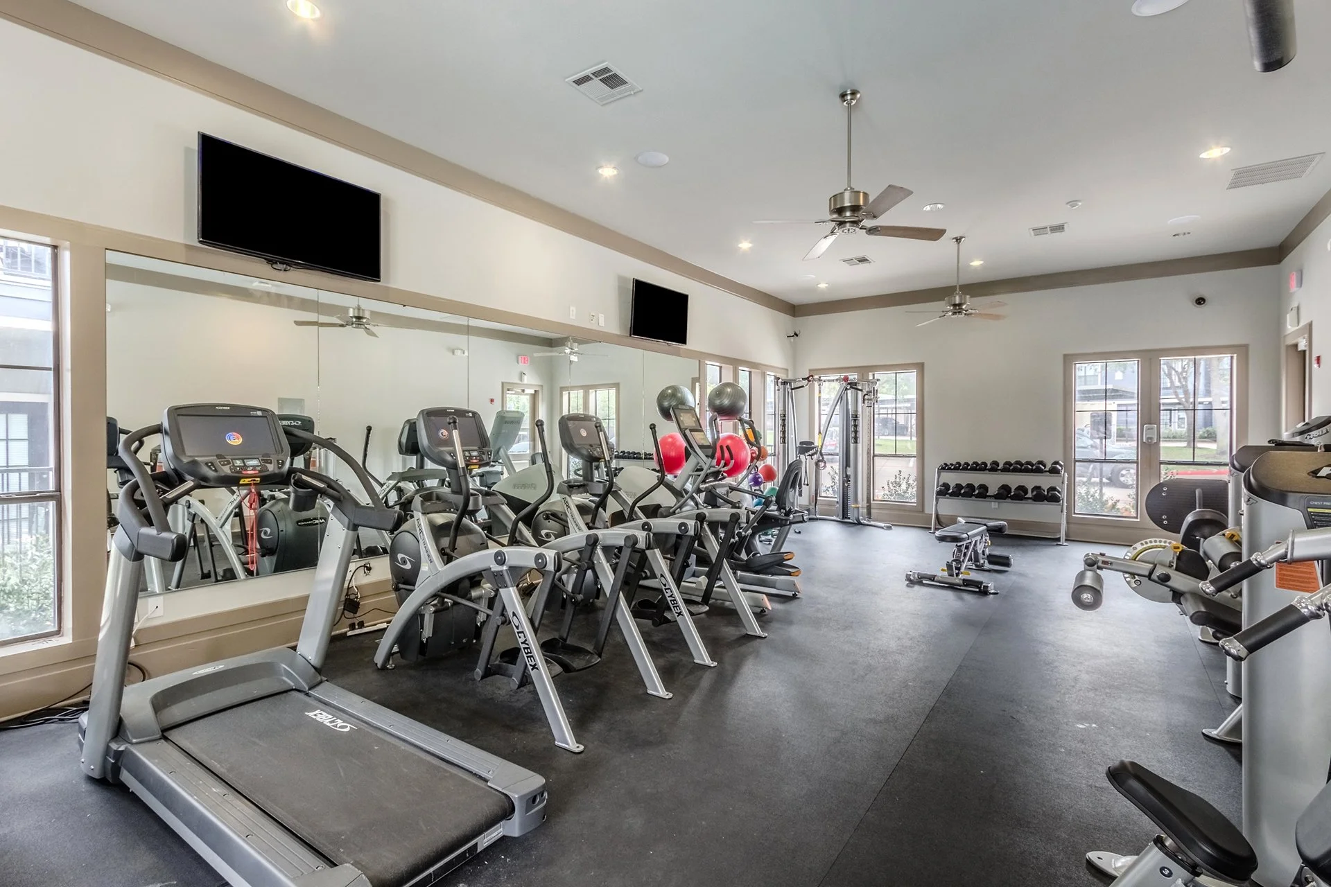 Indoor gym with treadmills, elliptical machines, dumbbells, and exercise balls, with a large wall mirror and wall-mounted TVs.