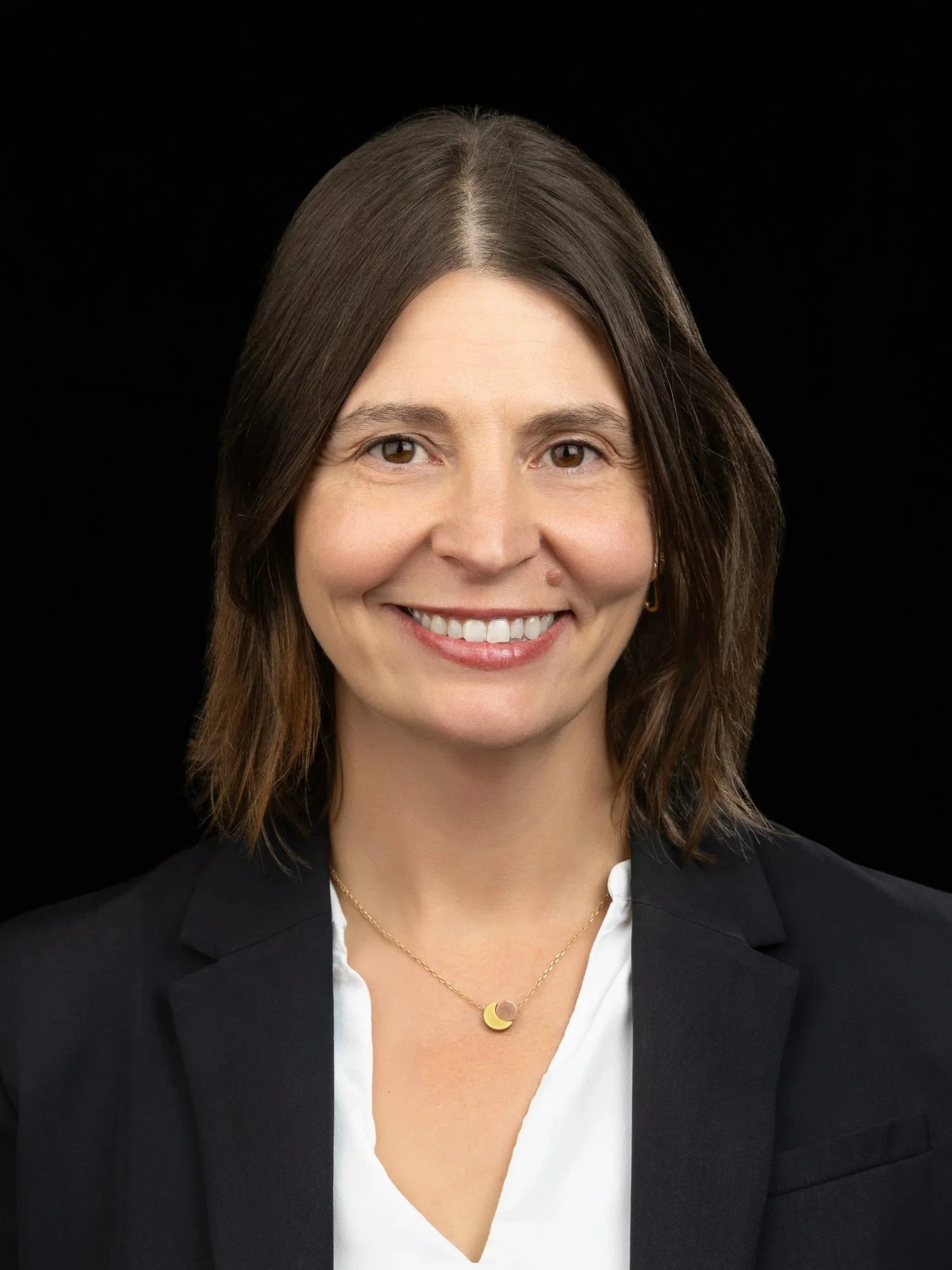 A woman with light brown hair, wearing a black blazer and a colorful beaded necklace, smiling against a dark brown background.