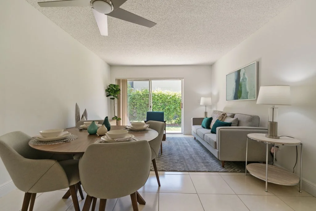 A living room with a dining area. The dining table is set with white plates, bowls, and decorative vases. The living room has a gray couch with teal and neutral pillows, a side lamp, and a framed artwork on the wall. A sliding glass door leads to an 