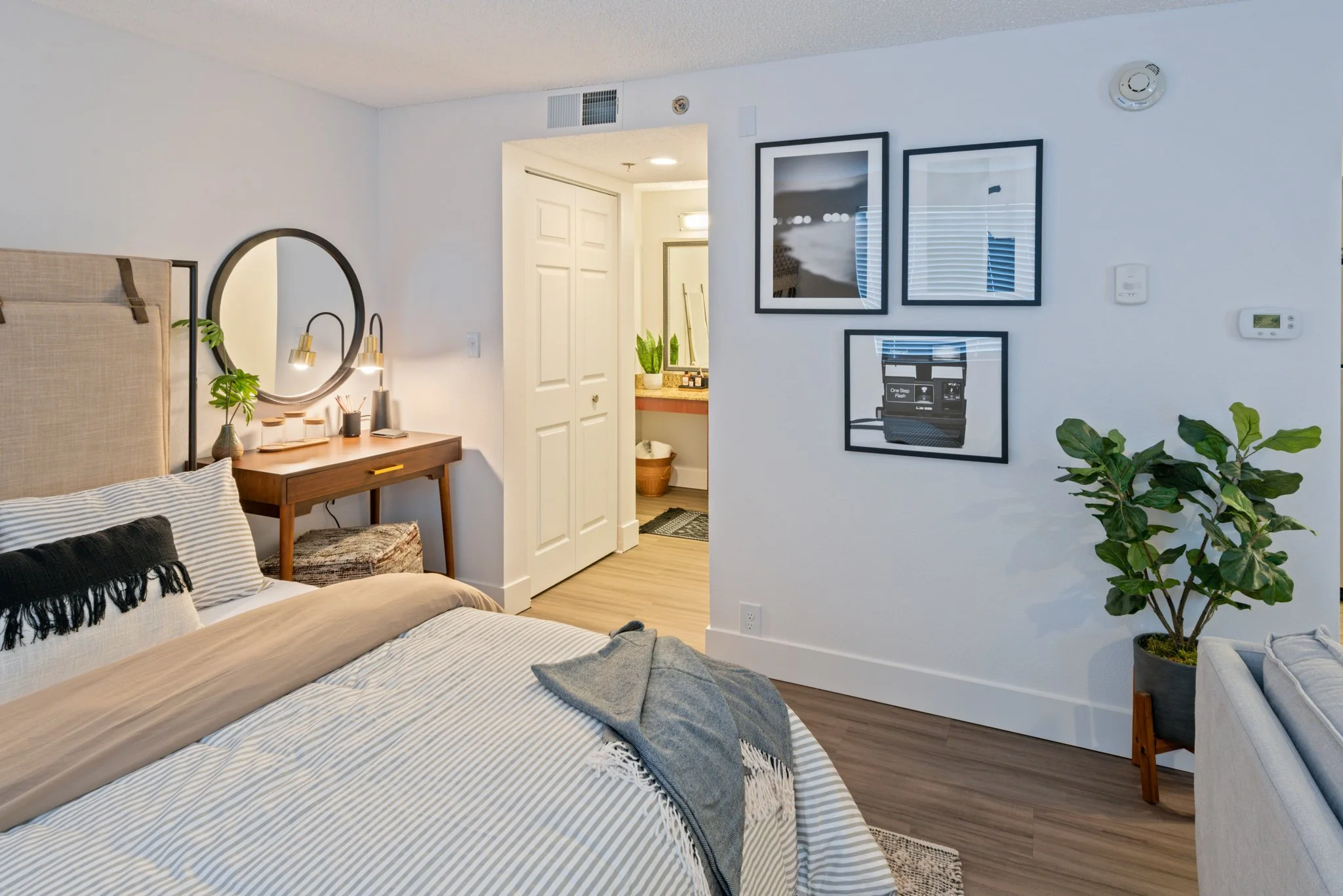 A modern bedroom with a bed, bedside table, mirror, framed photographs on the wall, a potted plant, and an entrance to a bathroom in the background.