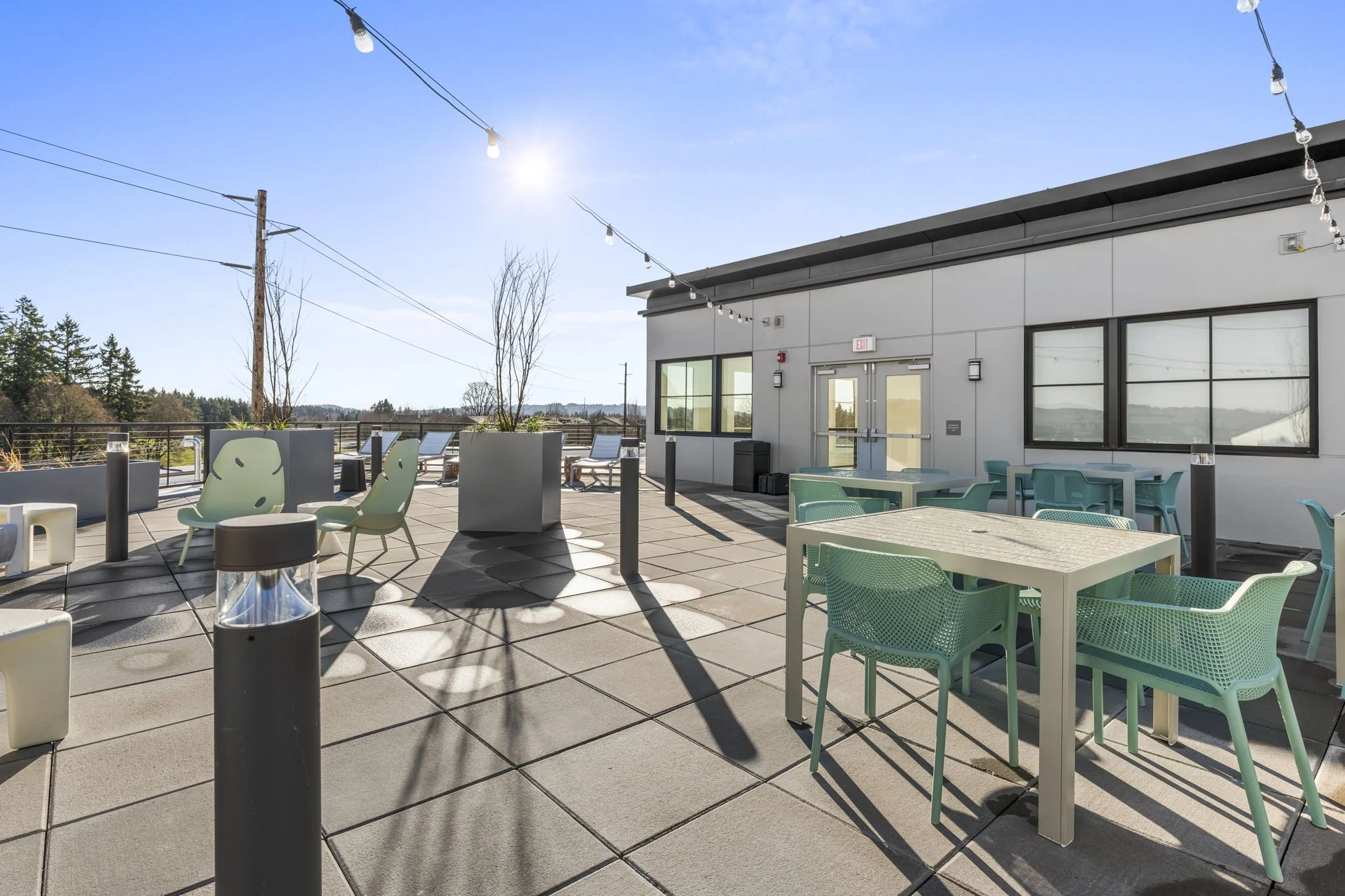 An outdoor rooftop patio with tables, chairs, and string lights, adjacent to a white building with large windows, under a clear blue sky with bright sunlight.
