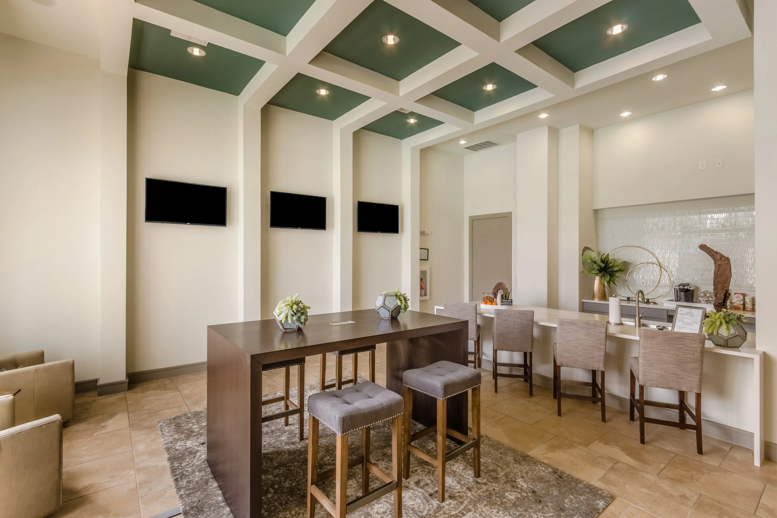 Modern lounge with high ceiling and recessed lighting, featuring wall-mounted TVs, a large wooden table with plants, and a bar area with bar stools and decorative items.