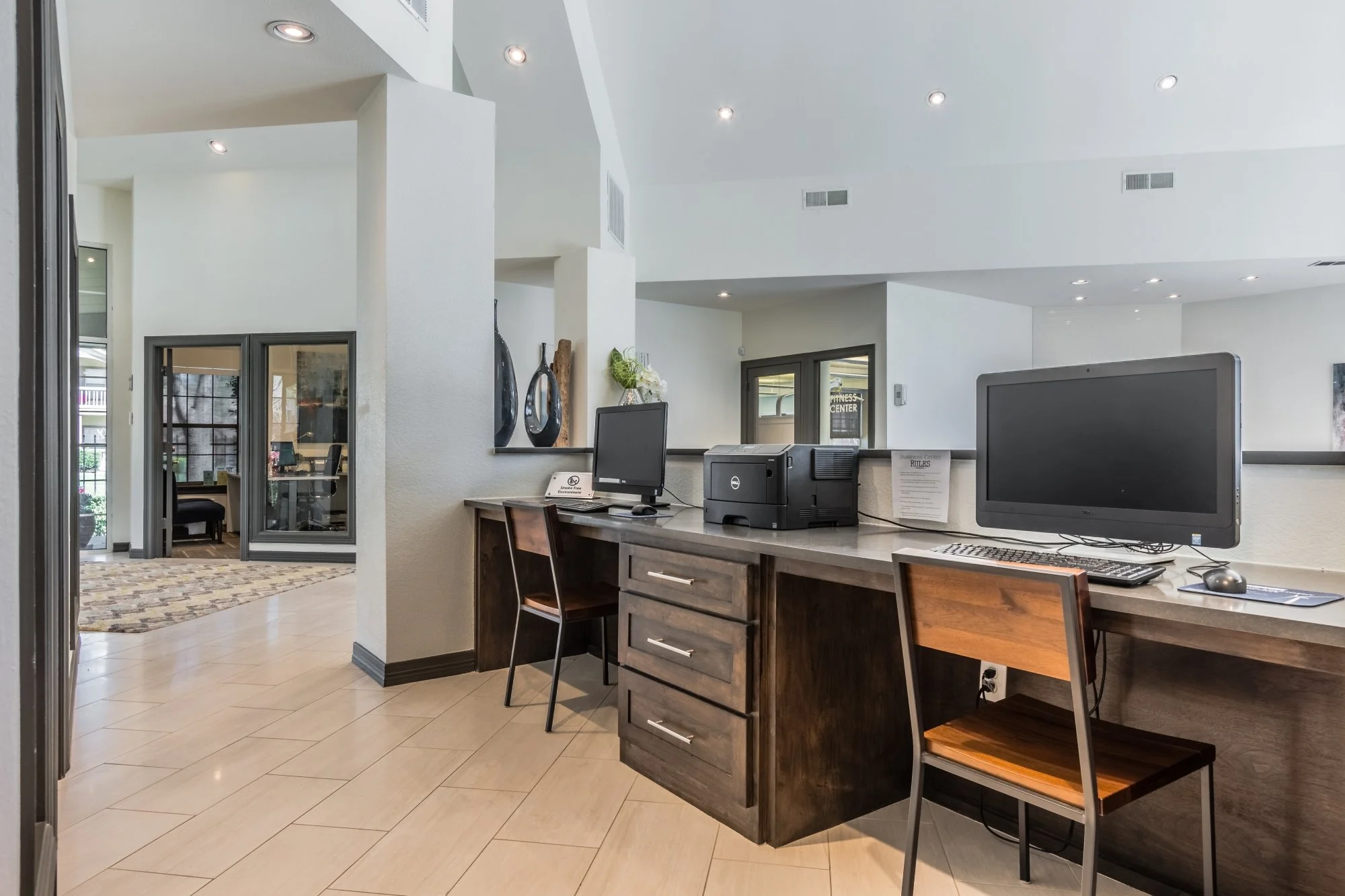 Hotel reception desk with two computer stations, chairs, a printer, and decorative vases in the background, with a sitting area visible through a glass window.