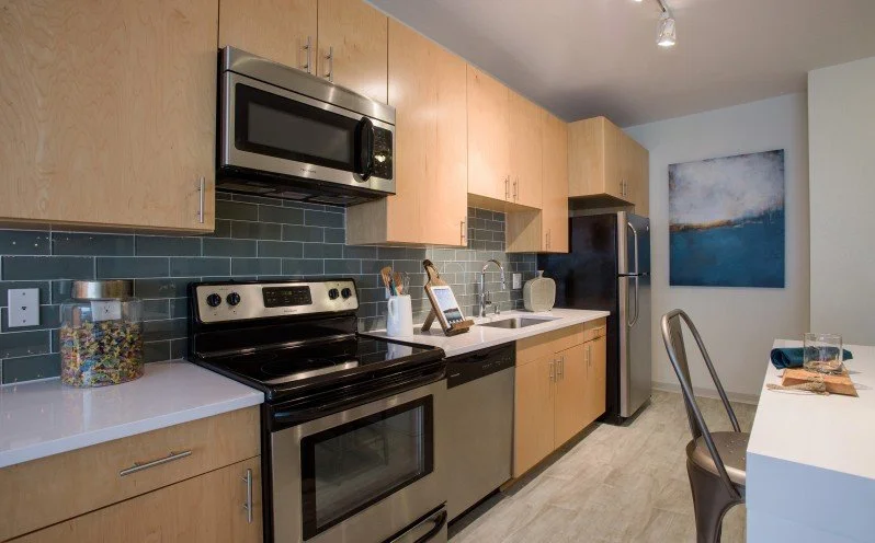 Modern kitchen with light wood cabinets, stainless steel appliances including microwave, oven, and refrigerator, dark gray subway tile backsplash, white countertop, and a dining table with chairs. There is a colorful jar on the counter and a blue abs