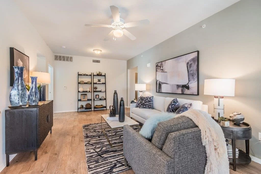 Living room with beige and gray sofas, abstract art on the wall, wooden flooring, and a black metal shelf with decorative items. There are two table lamps on side tables, a black and white patterned rug, and three tall black vases on a coffee table.