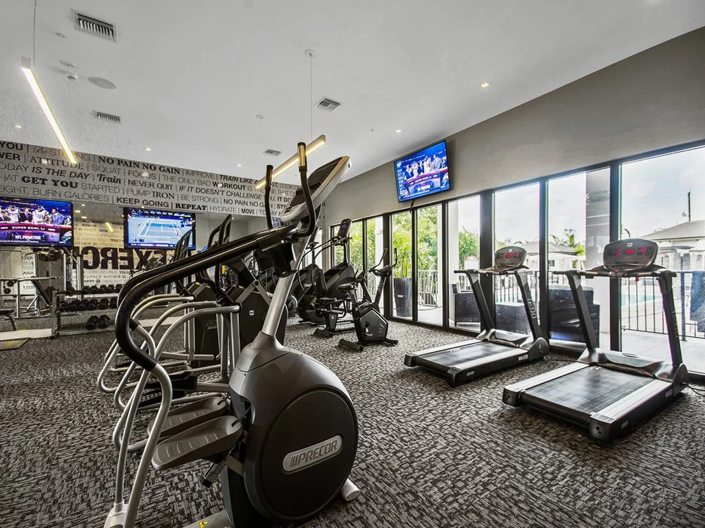 A fitness room with treadmills, elliptical machines, and exercise bikes, large windows, and wall-mounted televisions.