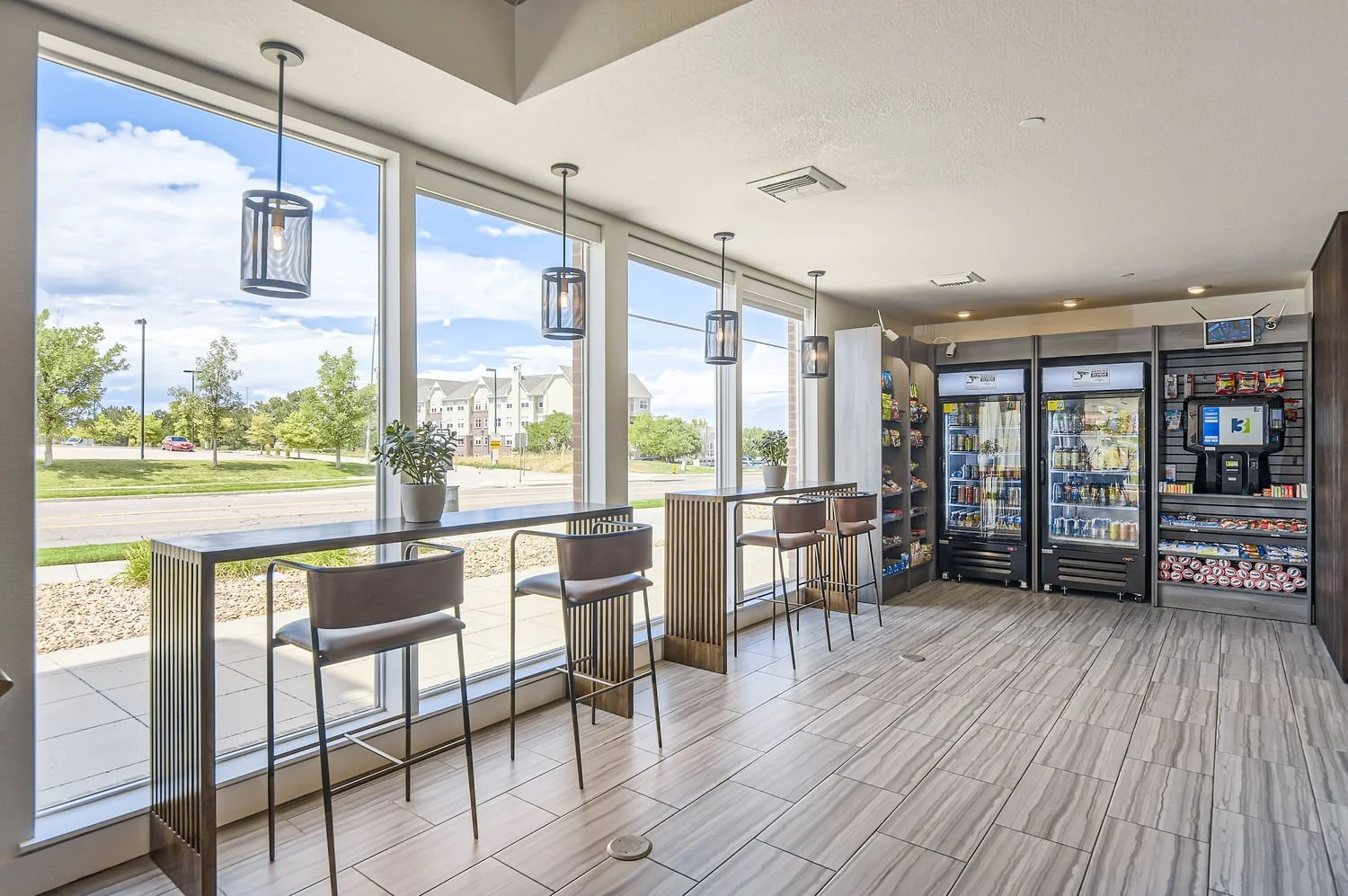 Interior of a modern convenience store or café with tall windows, pendant lighting, a high counter with chairs, and vending machines and snack shelves in the background.