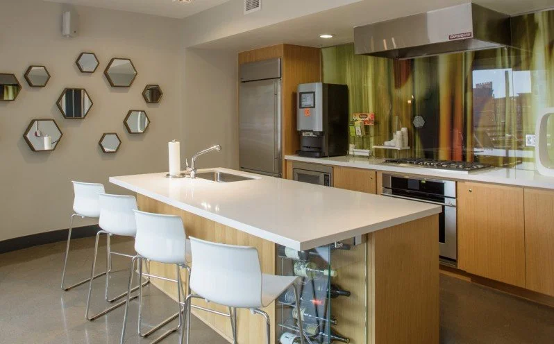Modern kitchen with a white countertop island, white bar stools, wooden cabinetry, a stainless steel oven, refrigerator, coffee machine, and decorative hexagon mirrors on the wall.