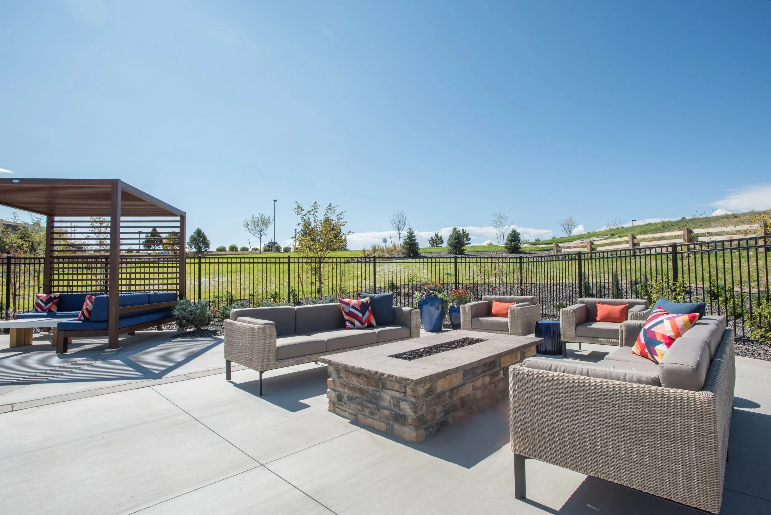 Outdoor patio seating area with wicker and fabric couches, chairs, and a fire pit, enclosed by a metal fence with a grassy hill and trees in the background under a clear blue sky.