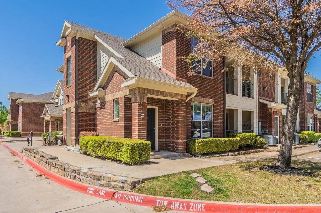 Exterior view of a brick apartment complex with a sidewalk, green bushes, and a tree in front. A red fire lane and no parking zone sign are visible in the foreground. Clear blue sky above.