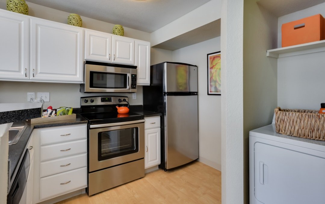 Kitchen with white cabinets, stainless steel microwave, stove with an orange kettle, and a refrigerator. There is a countertop with some items and artwork on the wall.
