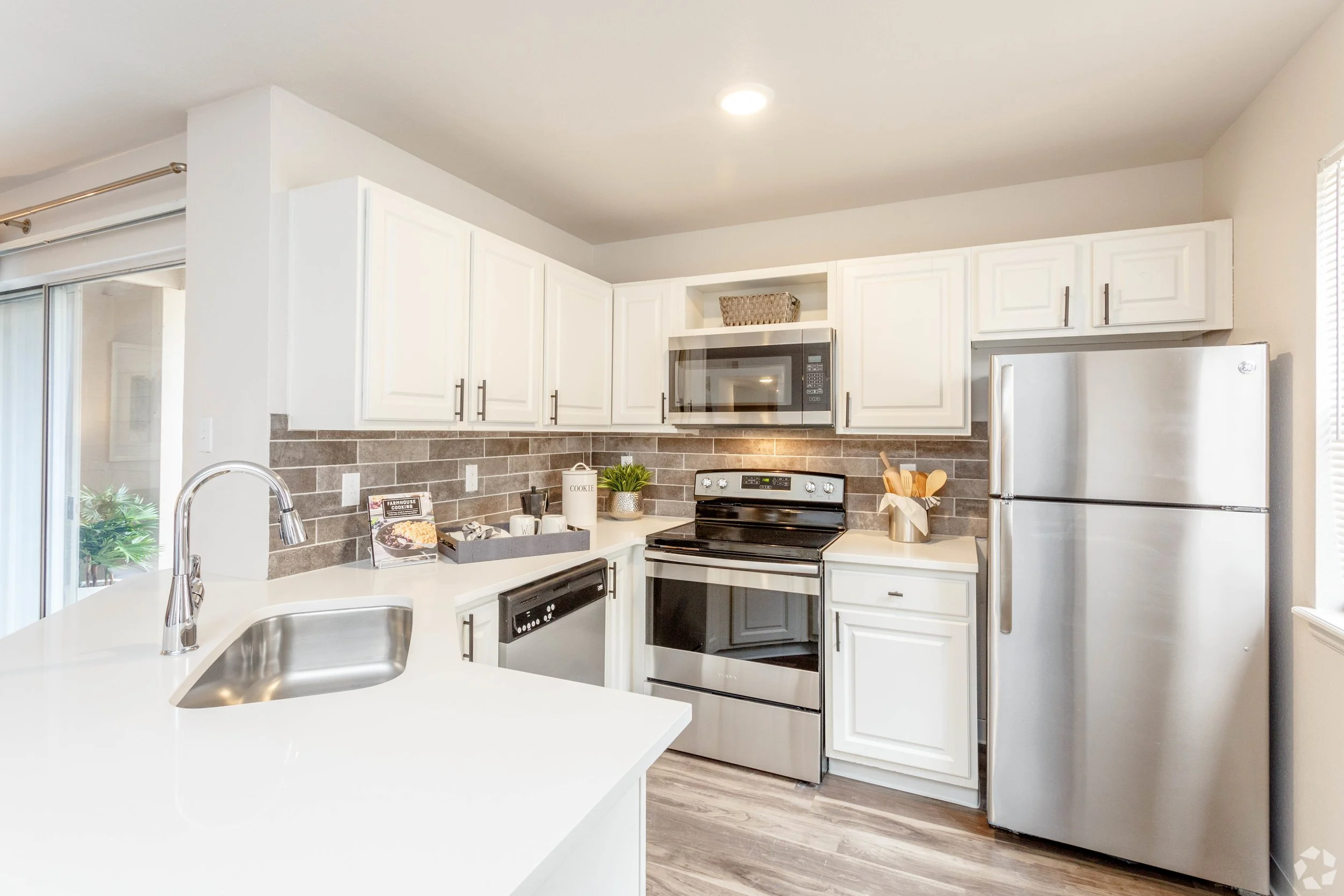 Modern kitchen with white cabinets, stainless steel appliances including a refrigerator, oven, microwave, and dishwasher. Features a gray brick backsplash, a white countertop, a sink, and decorative items like a plant, a cookie jar, and wooden utensi