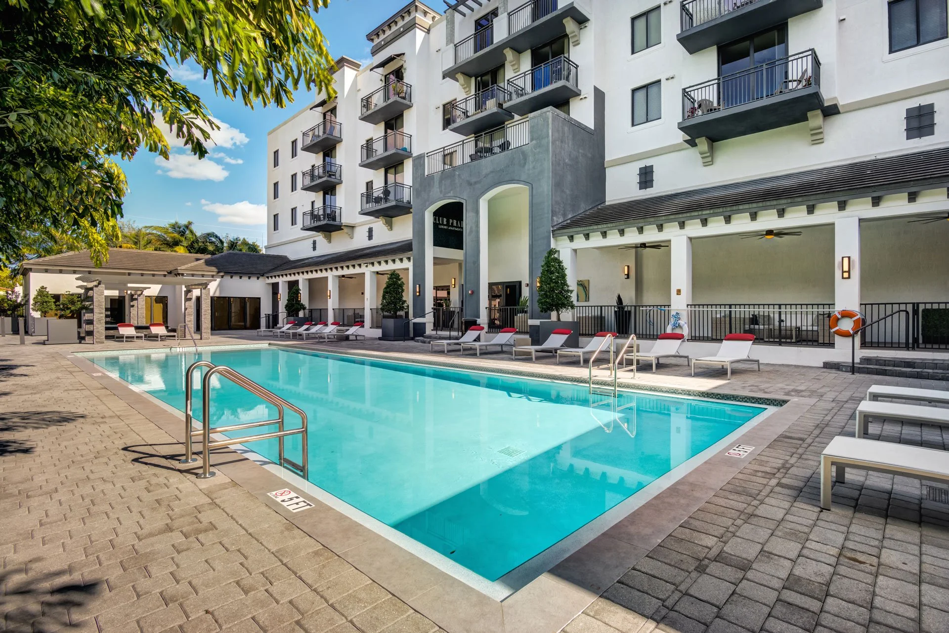 Empty outdoor swimming pool in a residential complex with lounge chairs, trees, and a modern apartment building in the background.