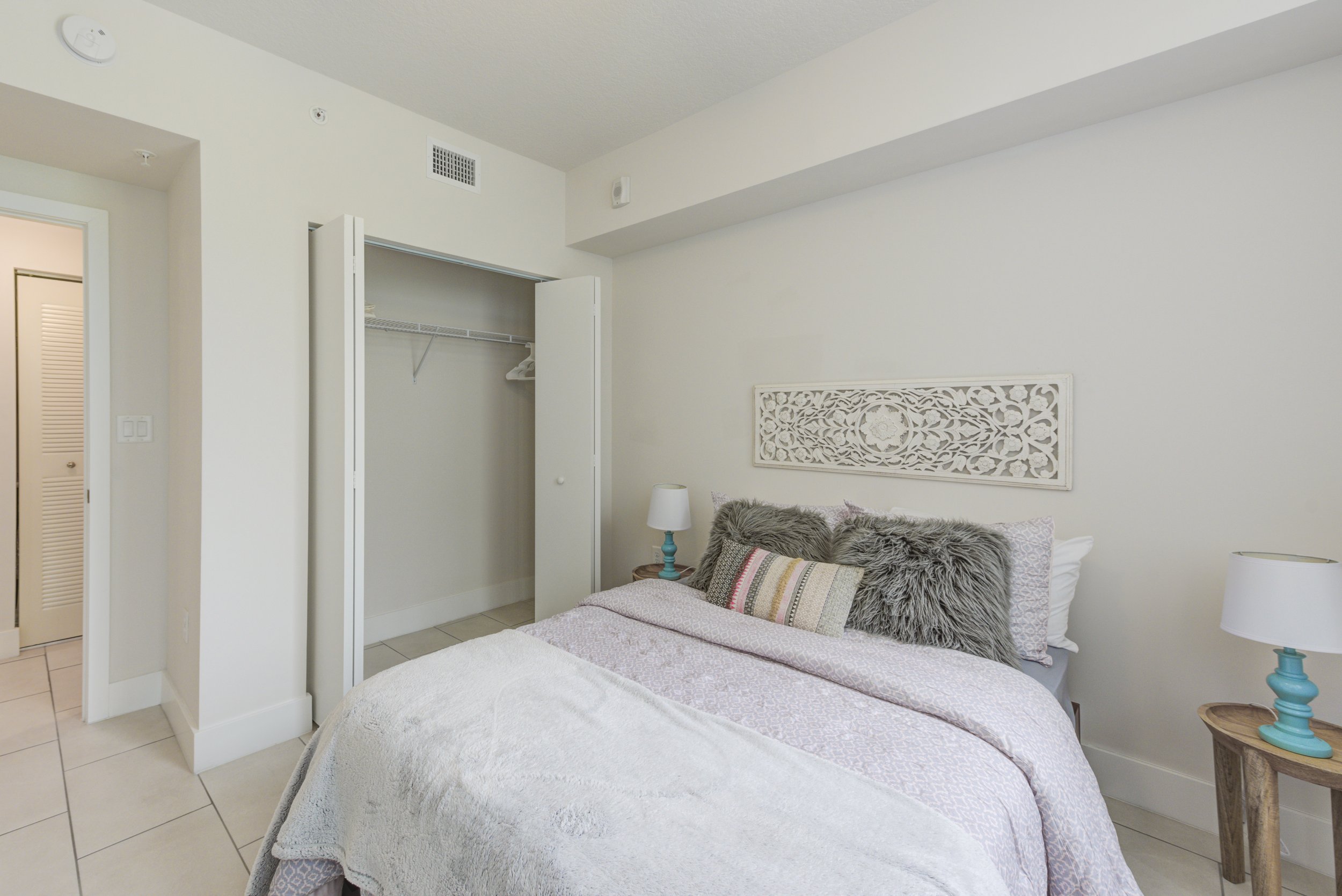 A bedroom with a bed covered with light-colored bedding, decorated with furry and striped pillows. There are small nightstands with lamps on each side of the bed. An open closet with a wire shelf and hangers is visible, and the walls are painted whit