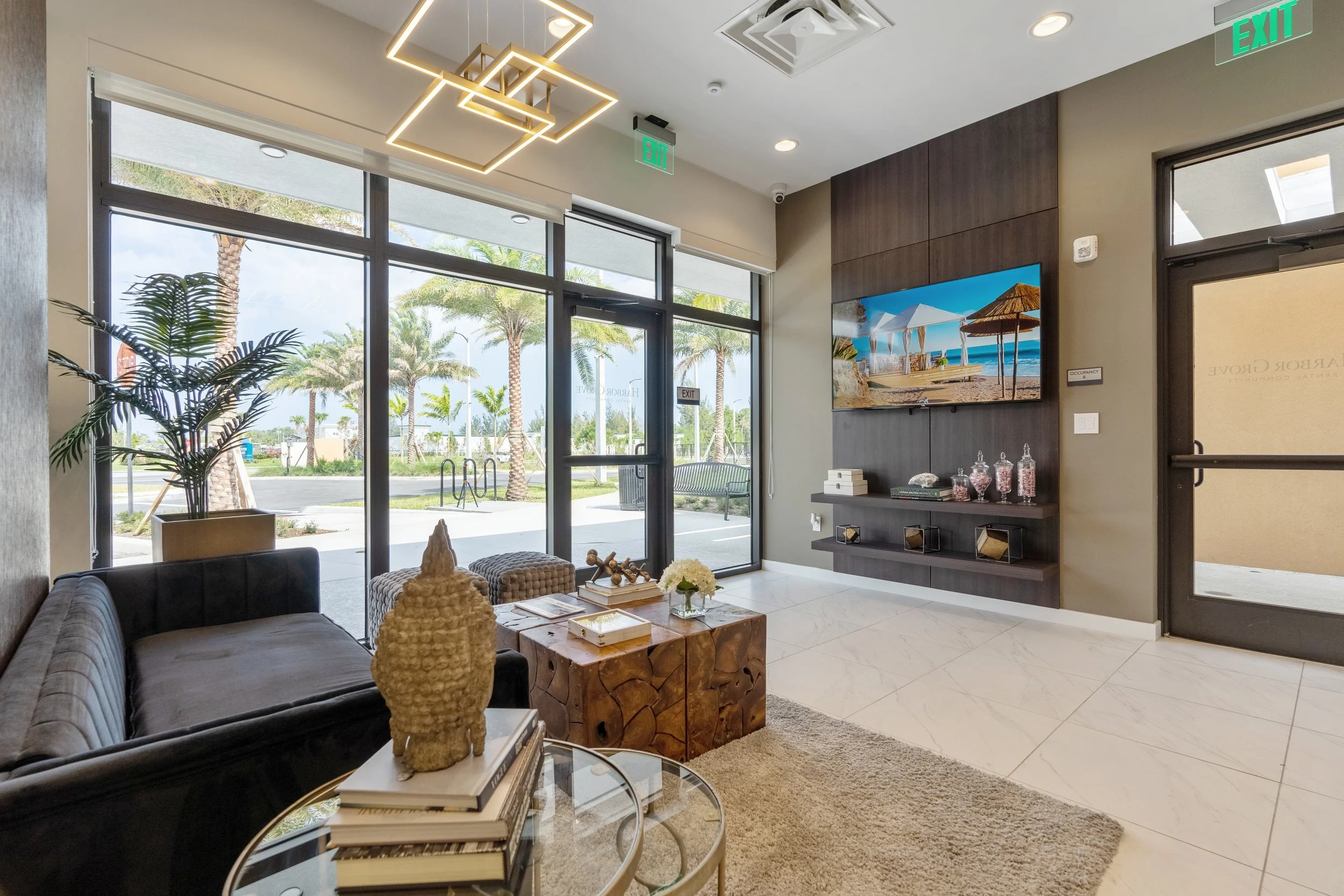 Modern hotel lobby with large glass windows showing palm trees outside. Inside features a black sofa, a glass coffee table with books and a decorative sculpture, a large flat-screen TV on a dark wood accent wall, and contemporary lighting fixtures.
